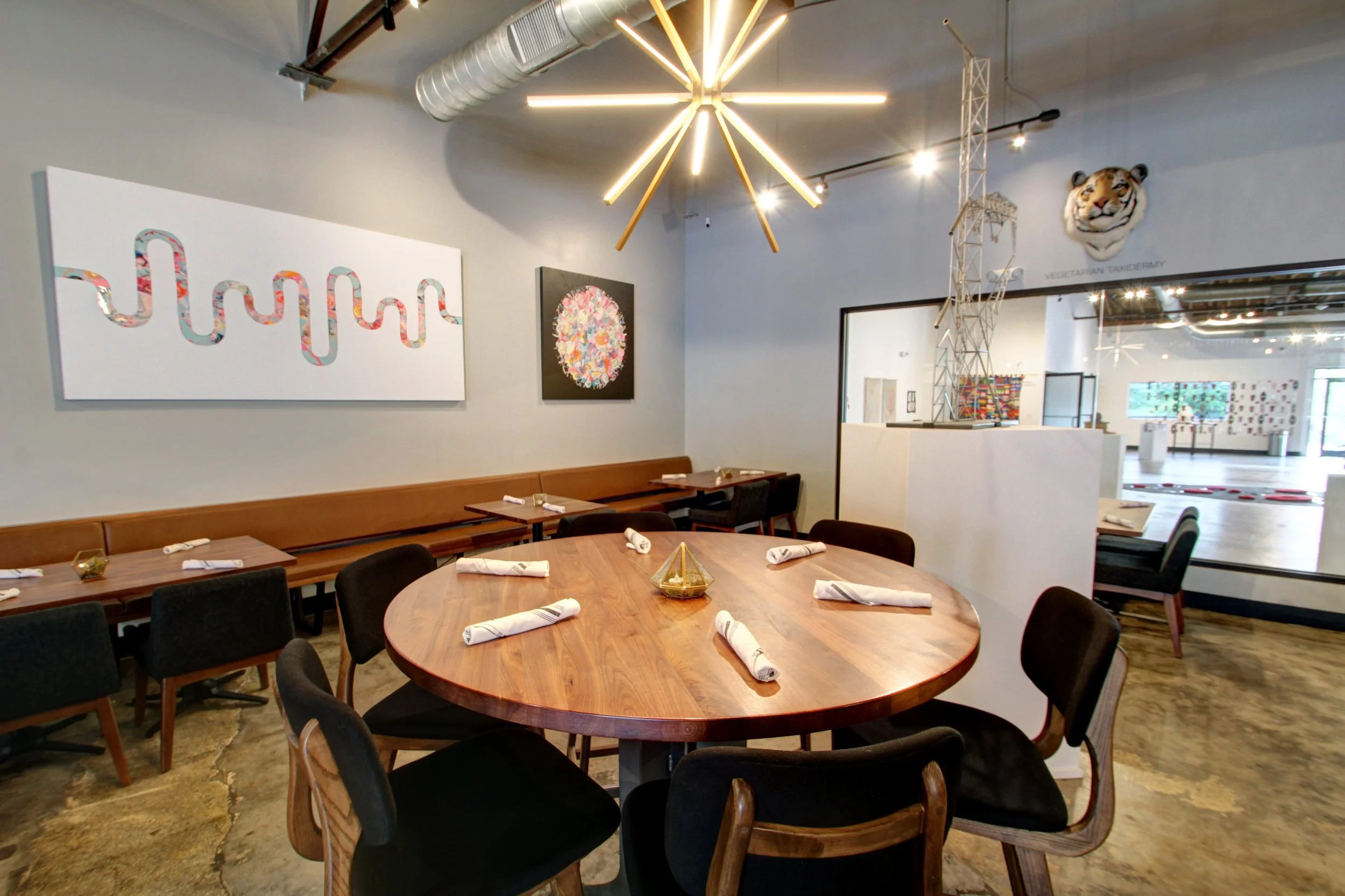 A modern dining area with a round wooden table set with white napkins, surrounded by black chairs, with artwork on the walls and contemporary lighting fixtures.