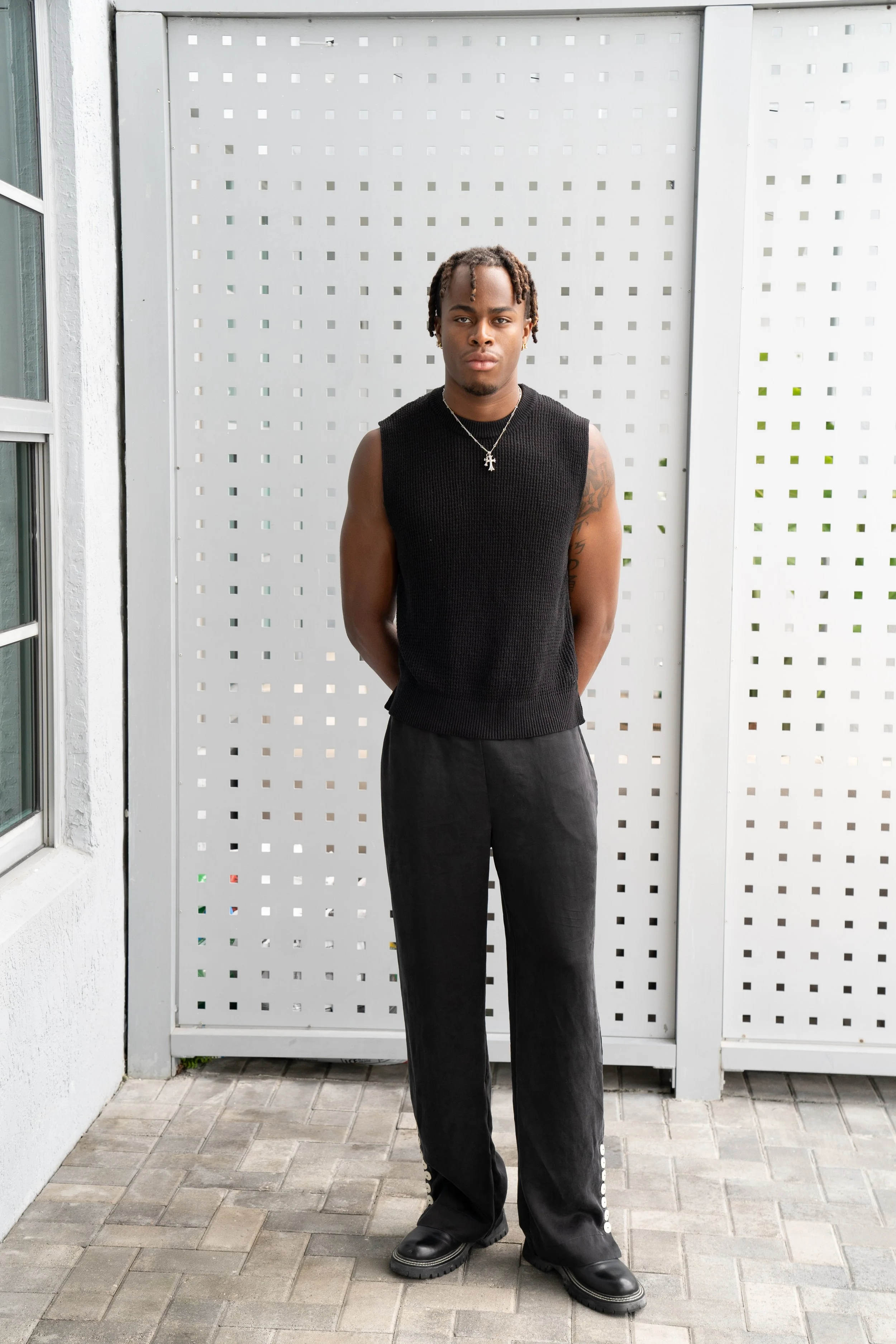 A young man with dreadlocks stands outdoors against a perforated white metal fence, wearing a sleeveless black knit vest, loose black pants, and black shoes, with a silver necklace featuring a cross pendant.