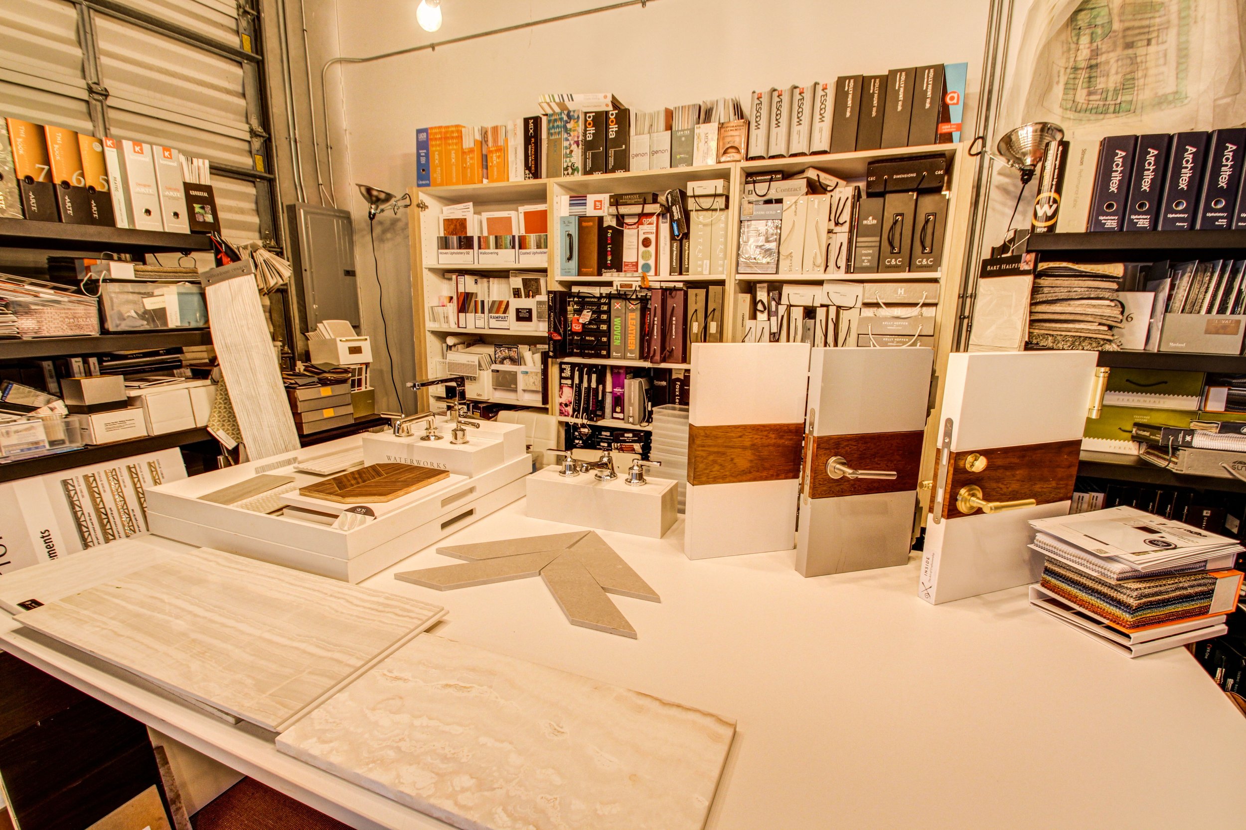Display of cabinet and door hardware samples, including door handles, knobs, and hinges, on a white tabletop in a hardware store or showroom with shelves filled with boxes and samples in the background.
