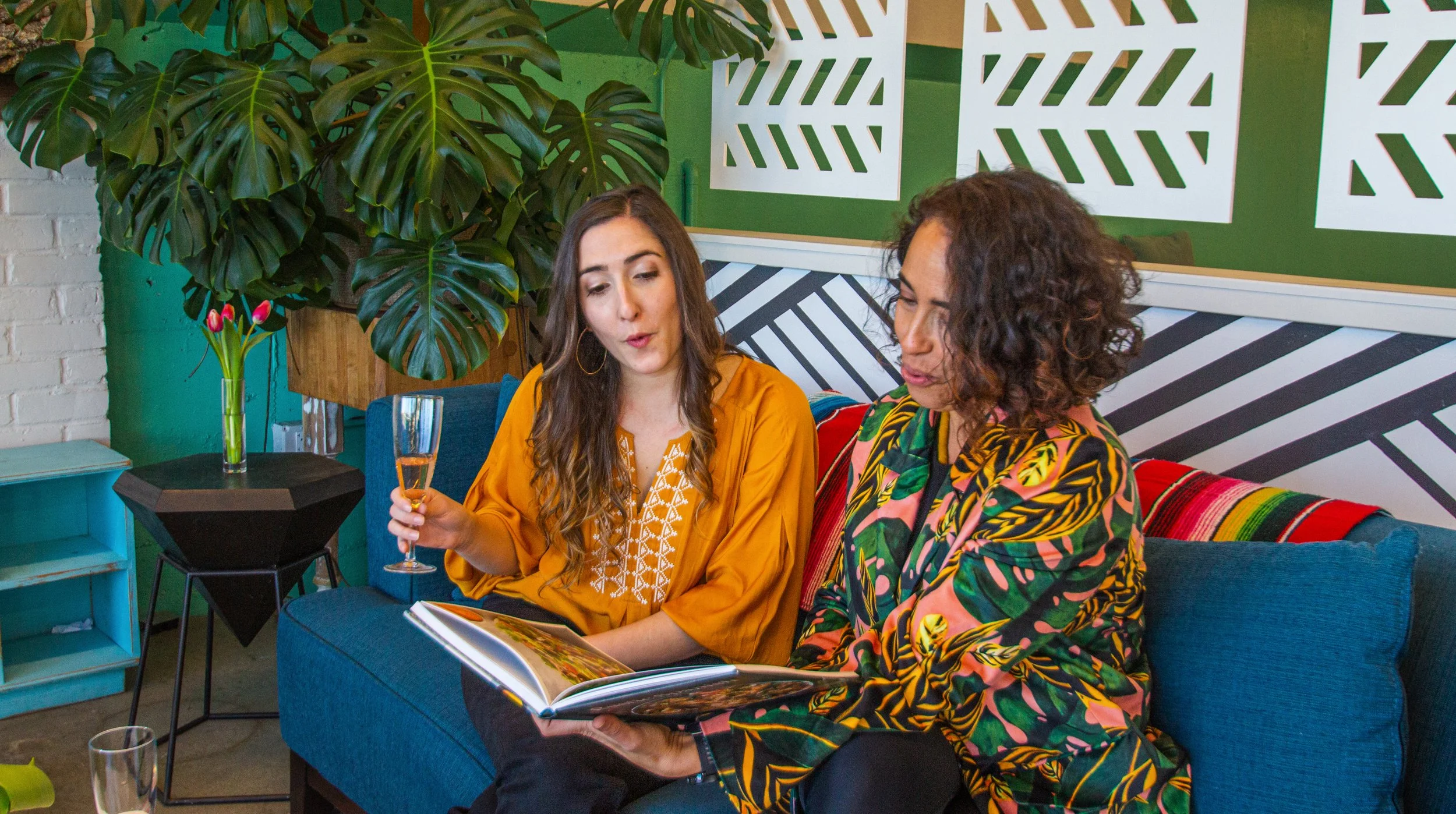Two women sitting on a blue couch in a colorful, tropical-themed room, looking at a menu, one holding a glass of what looks like champagne, with green plants and decorative patterned walls behind them.