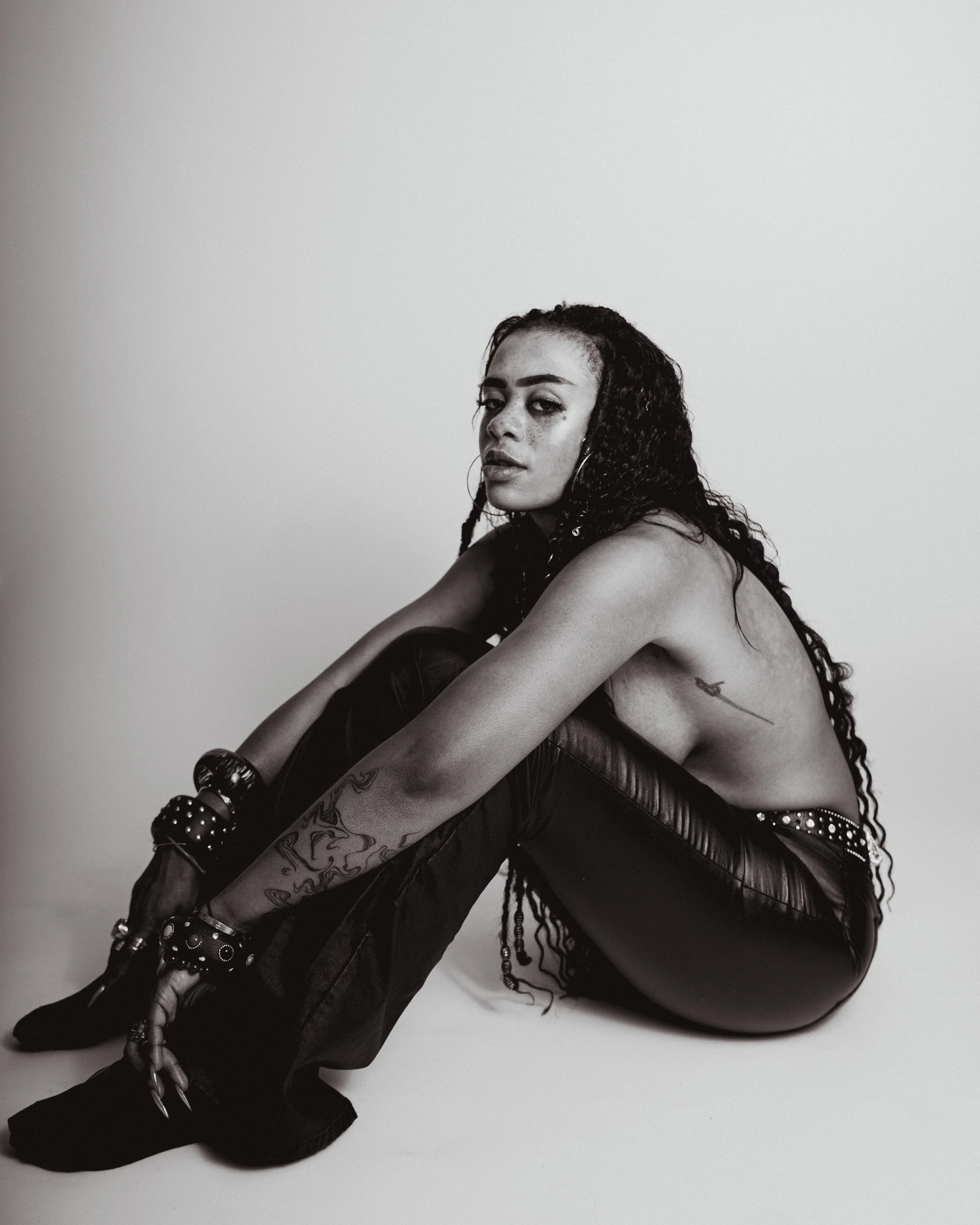 A black and white photo of a woman sitting on the floor with her knees drawn to her chest, wearing leather pants, and multiple bracelets. She has long curly hair and visible tattoos on her arm and side.