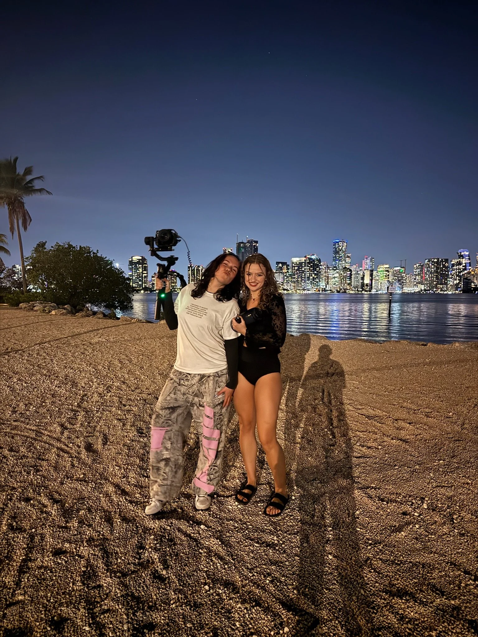 Two women posing together on a beach at night with a city skyline lit up in the background, one holding a camera on a tripod.