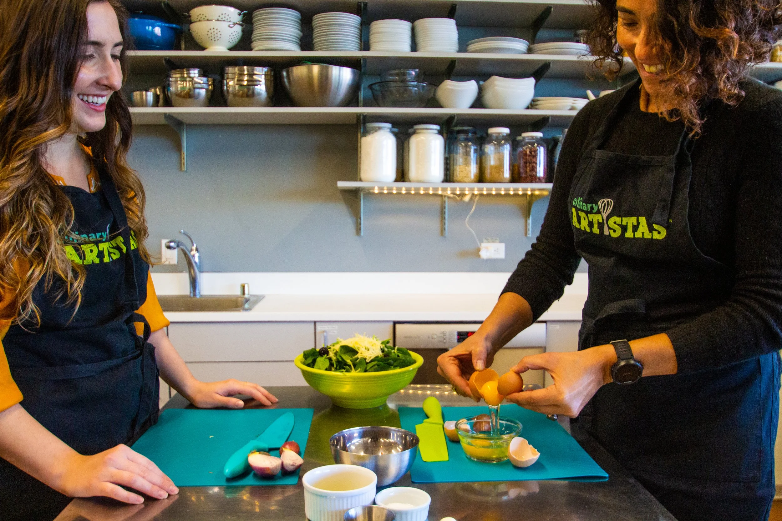 Two women cooking in a kitchen, cracking eggs into a bowl on a black countertop with a salad in a green bowl nearby.