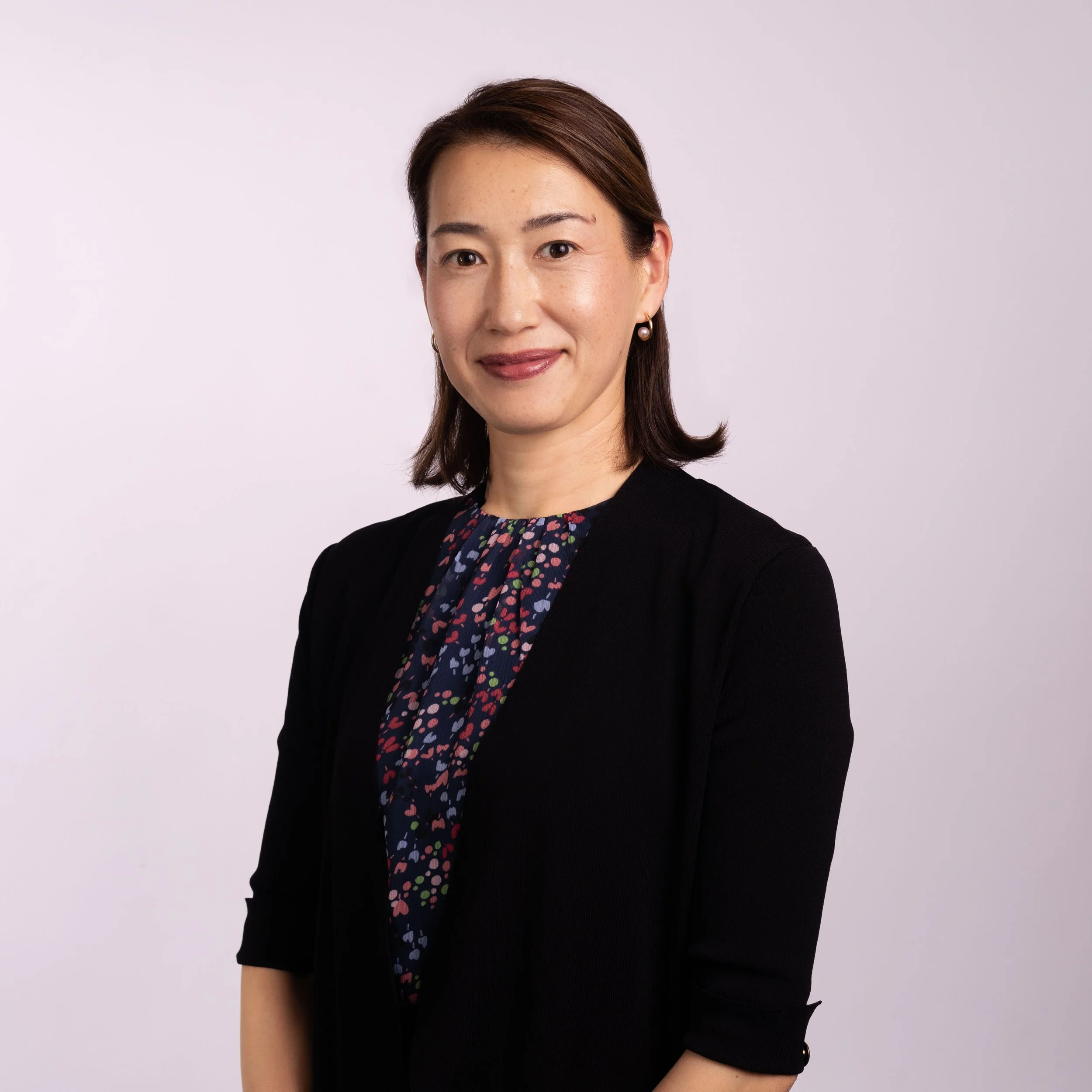 A professional woman with shoulder-length brown hair, wearing a black blazer over a colorful, patterned blouse, standing against a light gray background.