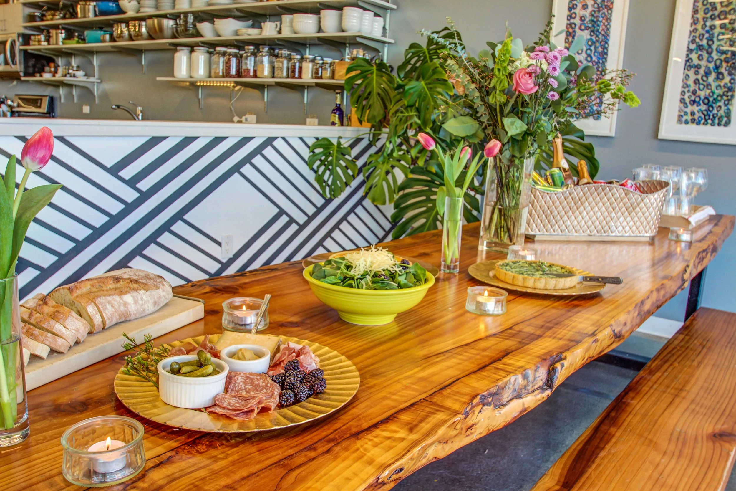 A wooden dining table set with food, flowers, and candles, in a cozy modern kitchen.