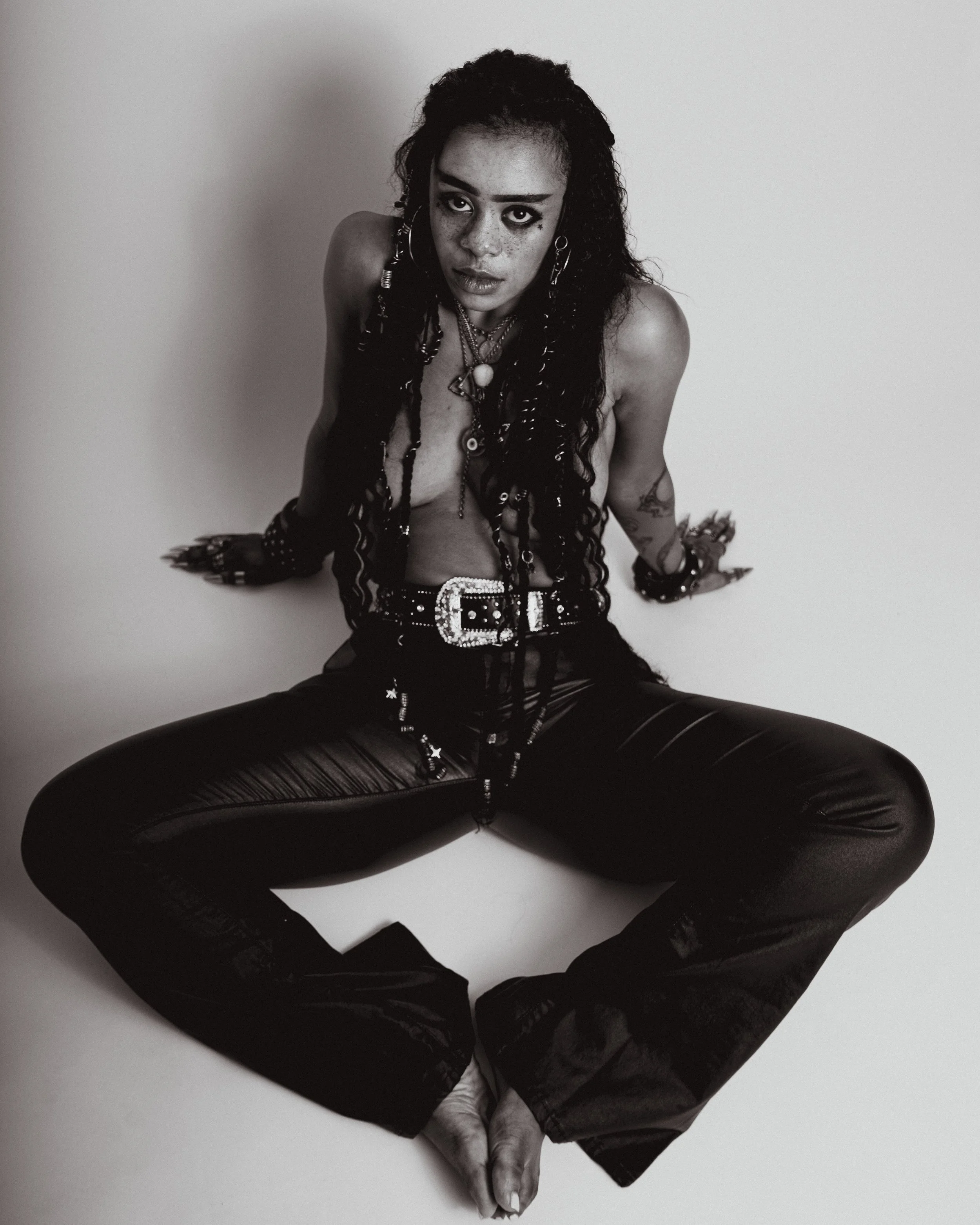 A woman with long, curly hair and earrings, sitting on the floor with her legs apart and looking directly at the camera. She is topless, wearing multiple necklaces, a studded belt, and shiny black pants.