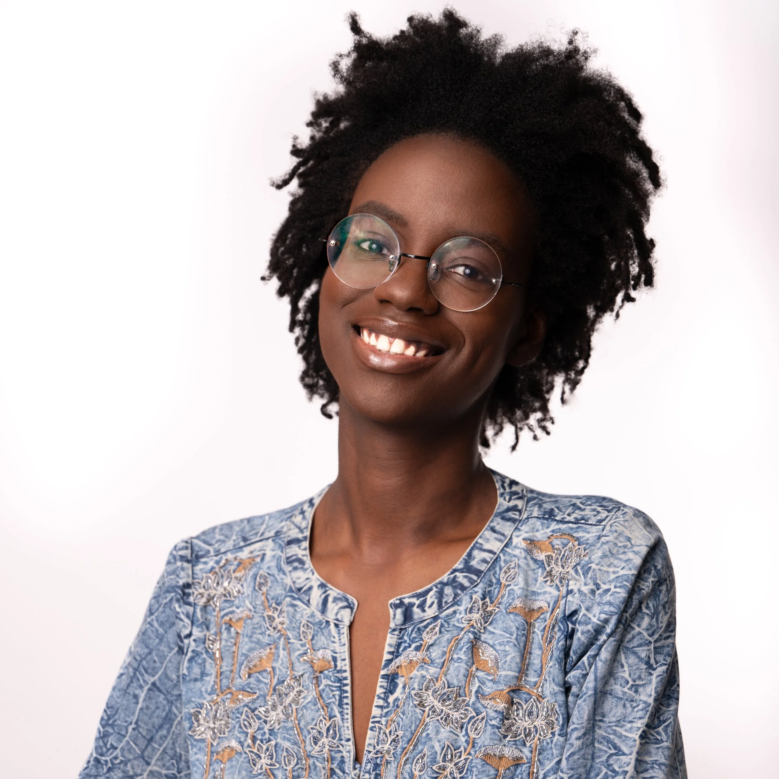 Portrait of a smiling woman with glasses, natural curly hair, wearing a blue embroidered blouse, against a plain white background.
