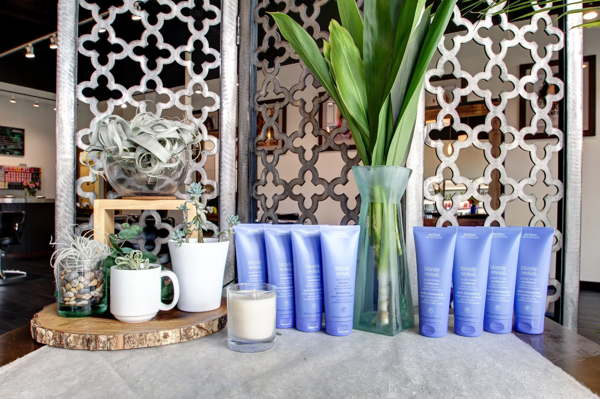 Various hair care products, including dozens of blue tubes of Aveda Blonde Revival, arranged on a table inside a salon or spa. The setup includes decorative plants and a candle, with a decorative partition in the background.