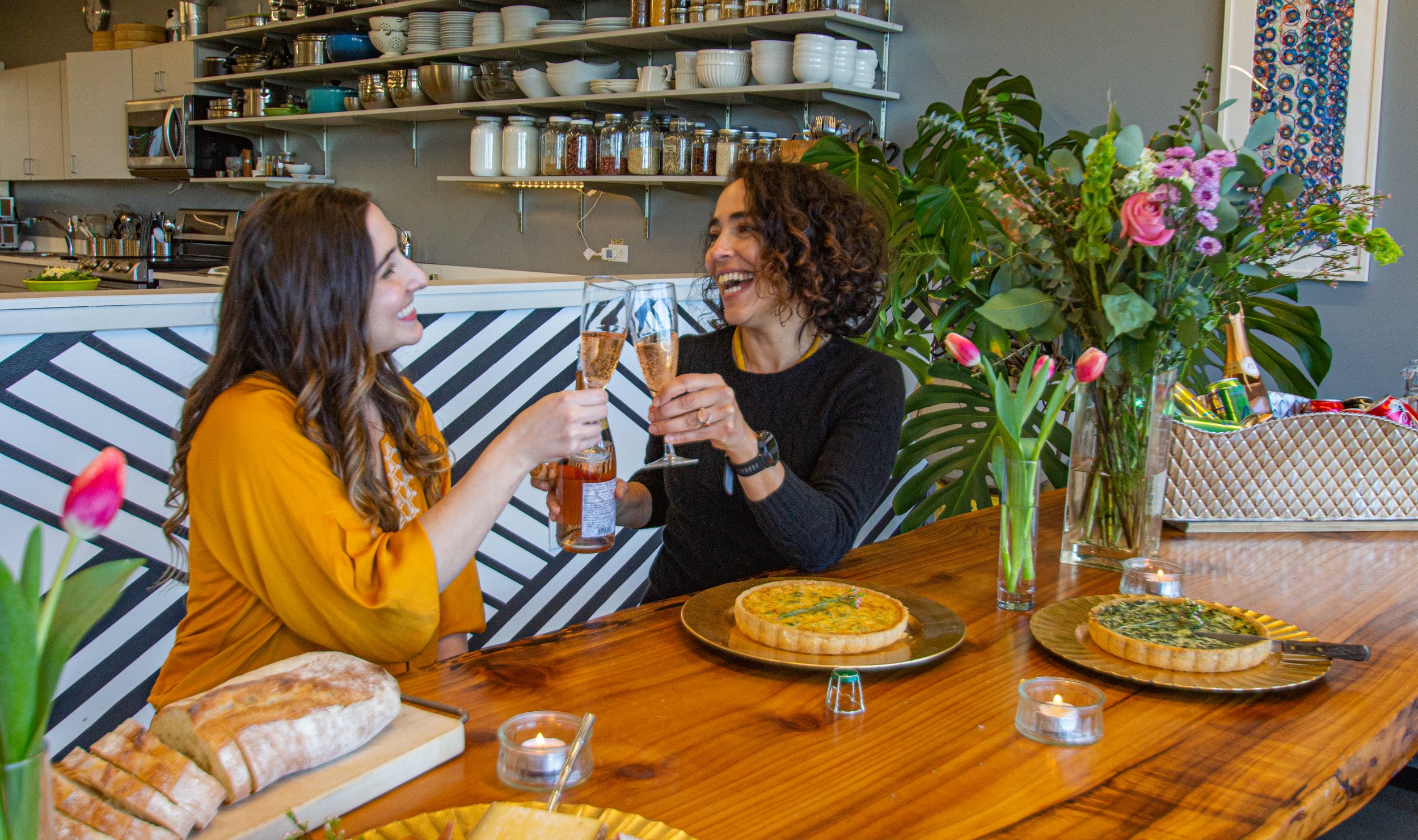 Two women toasting with glasses of rosé wine at a wooden dining table decorated with pink tulips and candles. The table has a loaf of bread, two pizzas, and a basket of snacks. The background features kitchen shelves with plates, jars, and kitchen eq