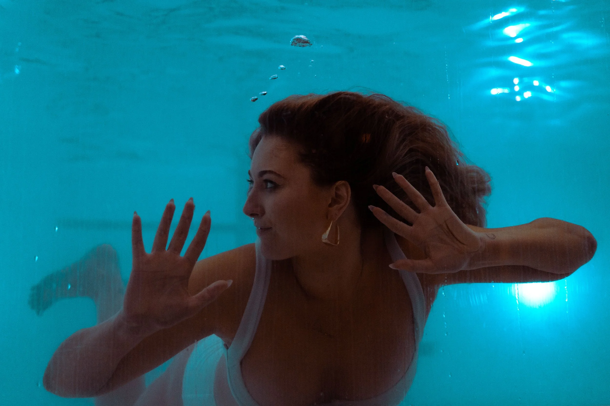 Woman with long hair and wearing earrings underwater, touching the glass of a swimming pool with both hands, facing sideways.