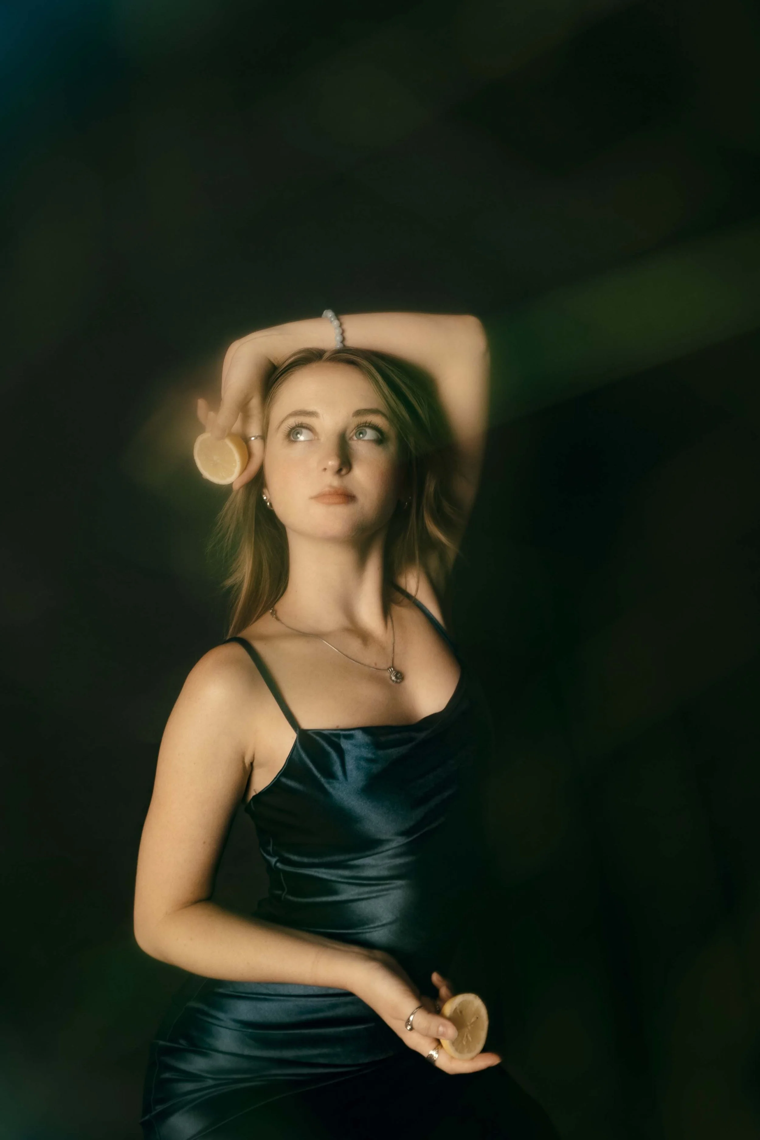 A young woman with shoulder-length hair in a black satin dress, holding a lemon slice, with one arm raised above her head, against a dark, blurred background.