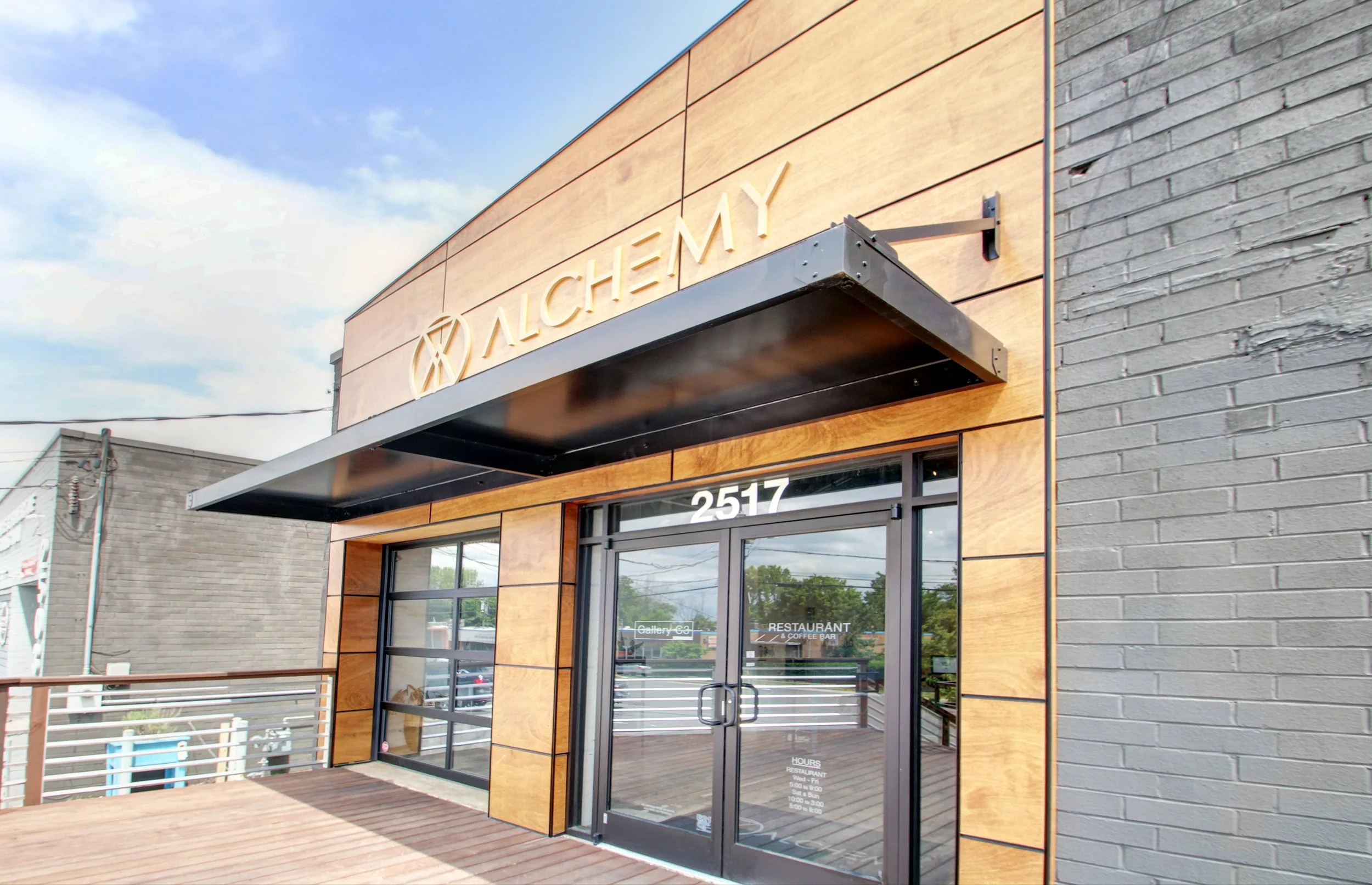 Exterior of a modern restaurant called Alchemy with large windows, wooden paneling, and a metal awning over the entrance, located at 2517.