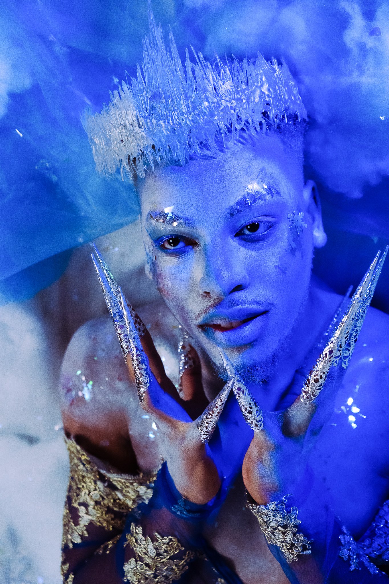 A person with elaborate, shiny costumes and makeup, adorned with metallic and glittery accessories, posing with hands near face under blue lighting.