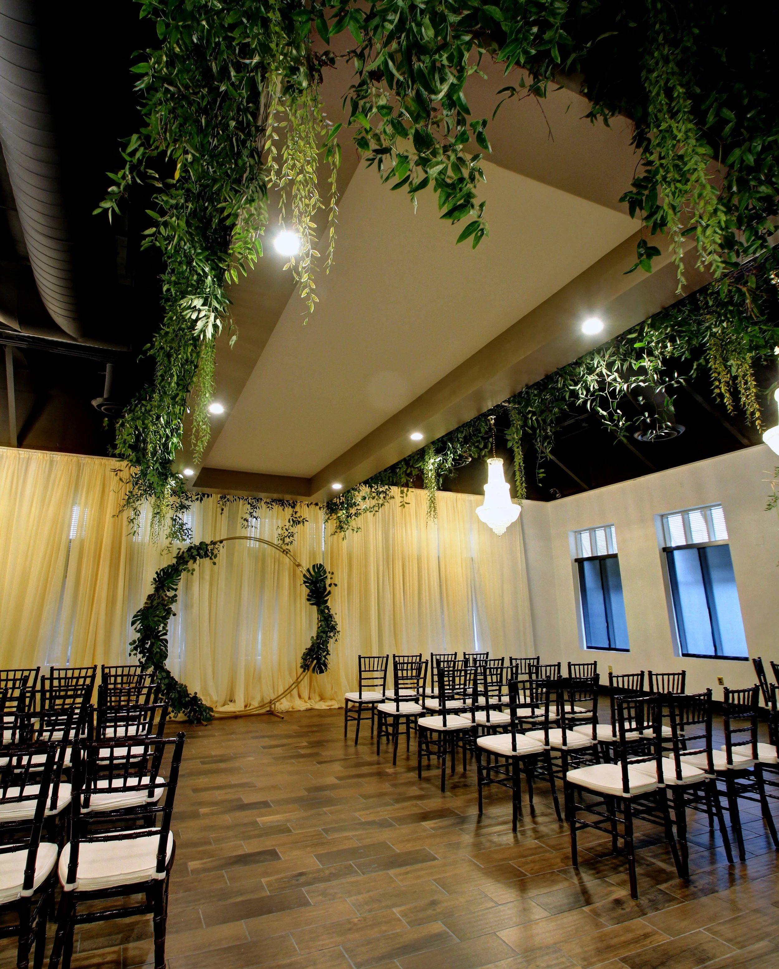A decorated wedding ceremony space with a circular floral arch, cream curtains, wooden chairs with white cushions, hanging chandeliers, greenery on the ceiling, and windows on the side wall.