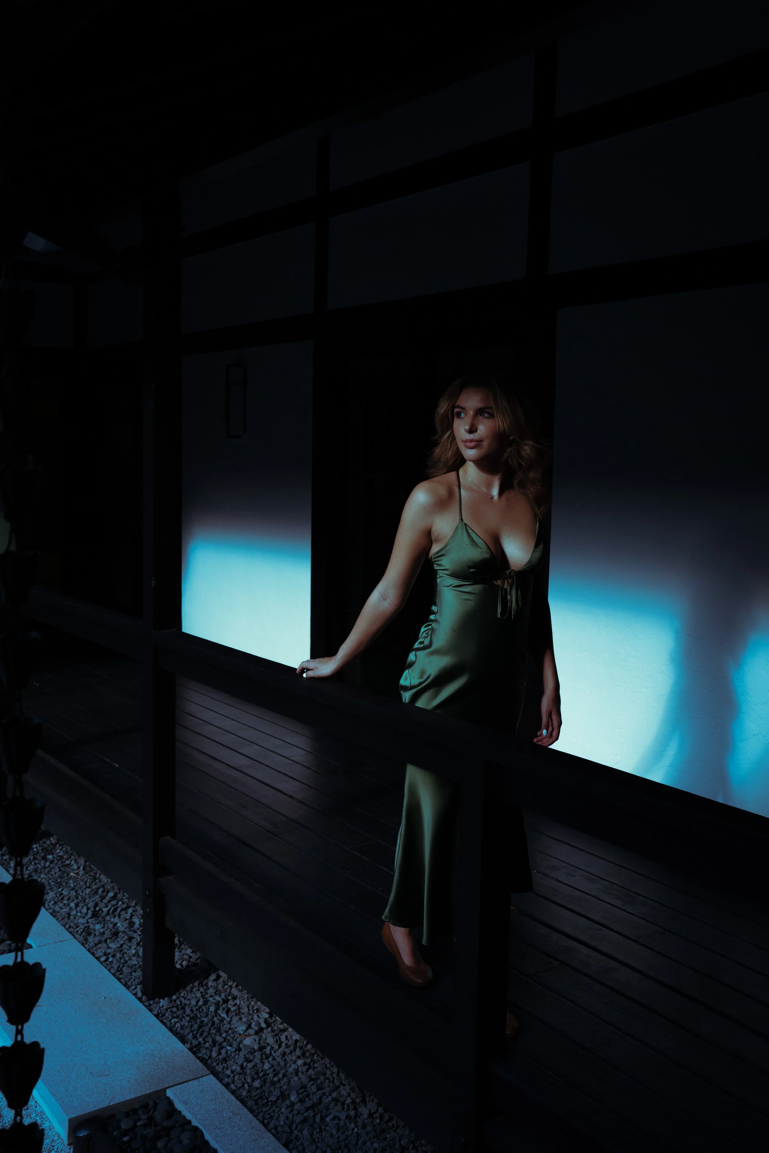 A woman in an olive green satin dress with spaghetti straps standing outdoors at night on a wooden porch, illuminated by blue light.