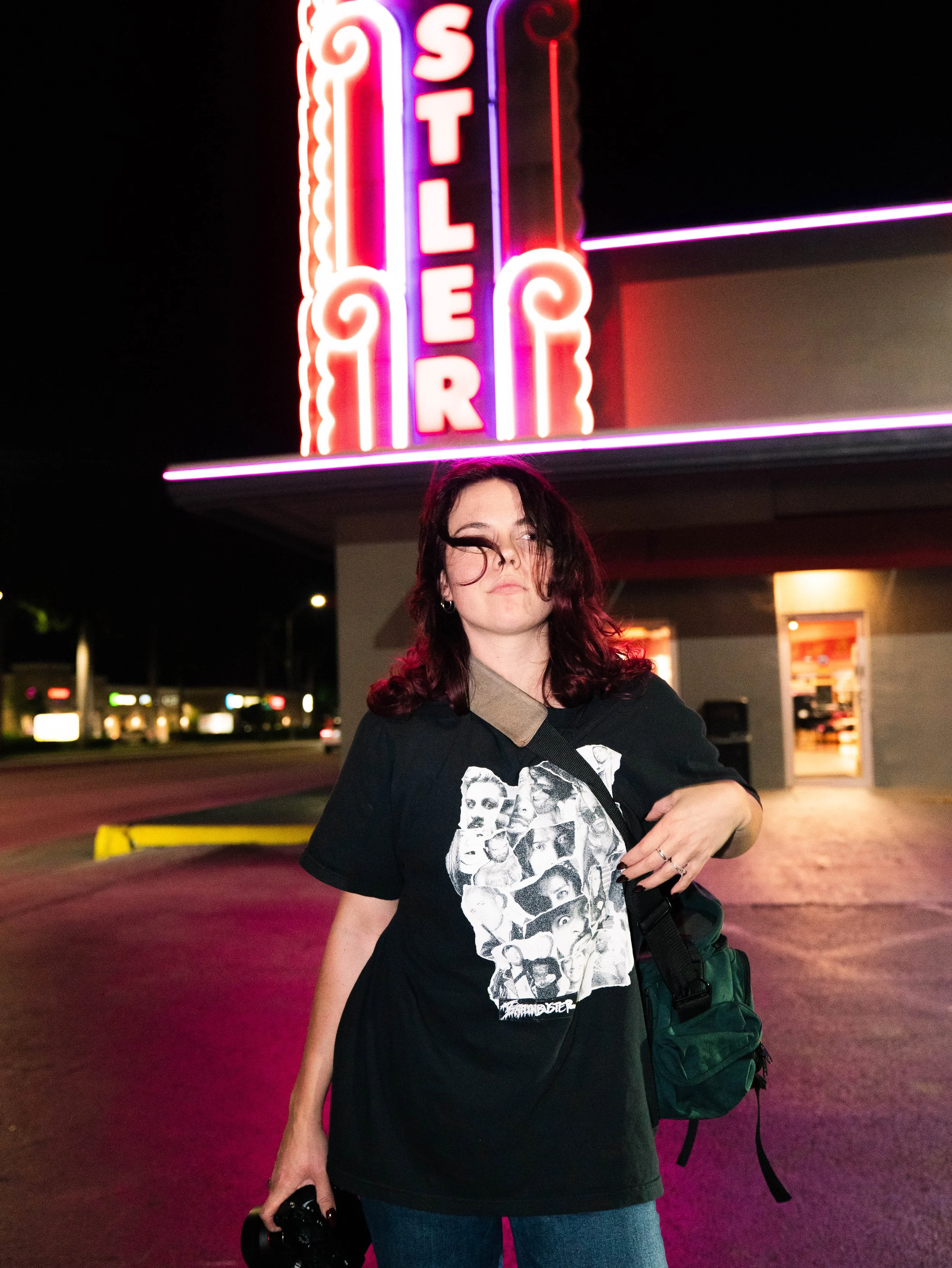 A woman standing at night in front of a neon sign that reads 'STH LNER'. She is wearing a graphic black T-shirt featuring faces, jeans, and a crossbody bag, holding a camera in her left hand.