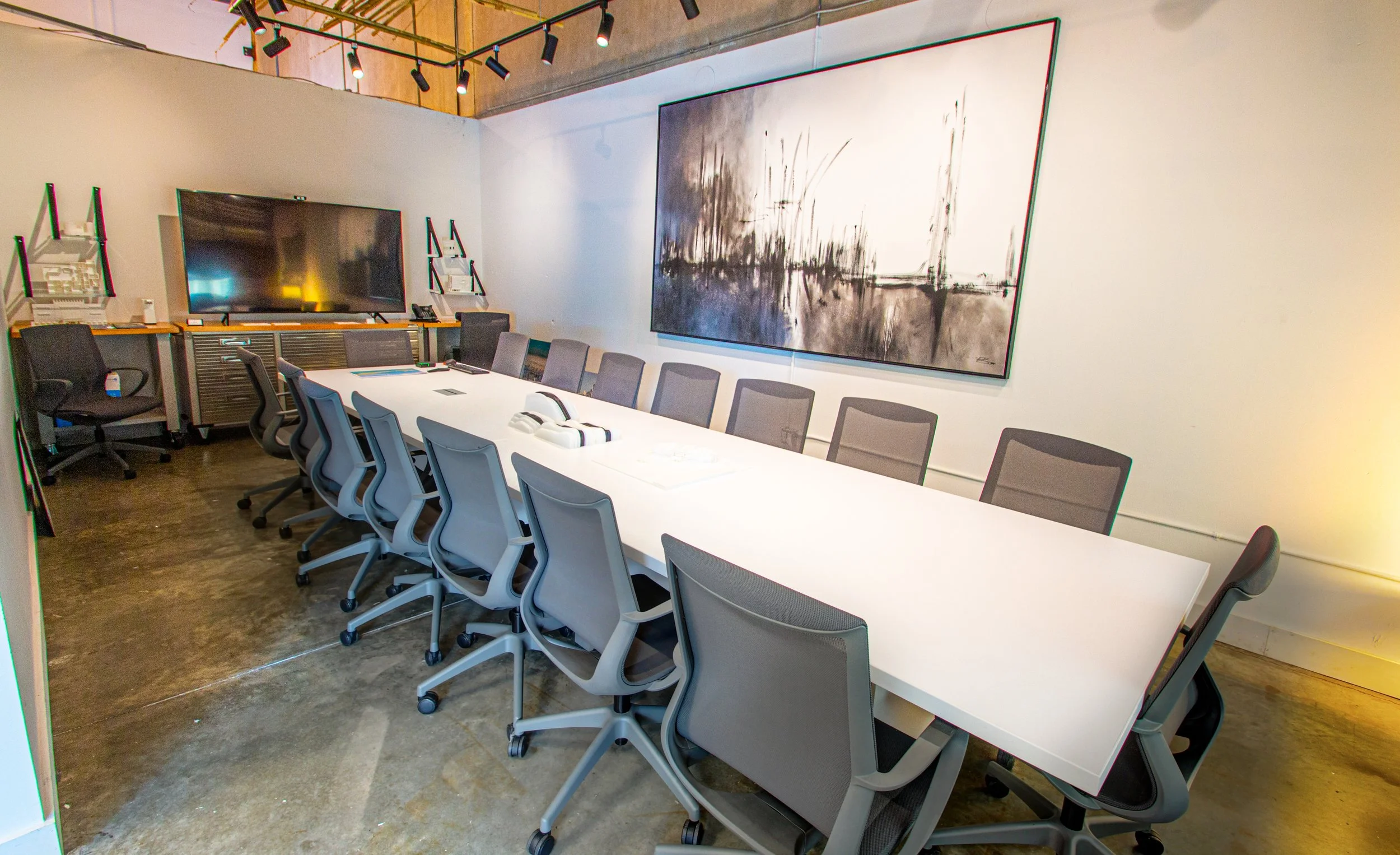 Modern conference room with a long white table, multiple ergonomic chairs, large wall-mounted display, abstract black and white artwork, and industrial-style ceiling lighting.