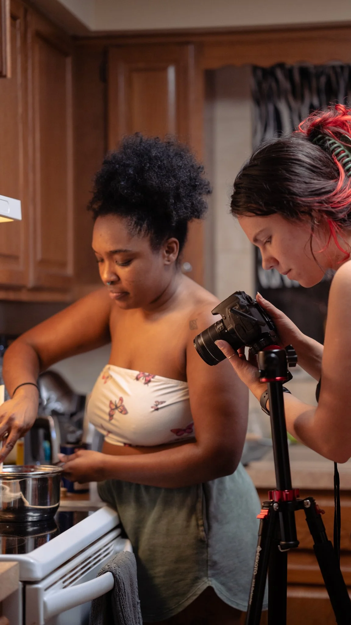 Two women are in a kitchen, one with dark curly hair wearing a floral crop top and gray sweatpants, and the other with red-tipped hair holding a camera on a tripod, taking photos or recording.