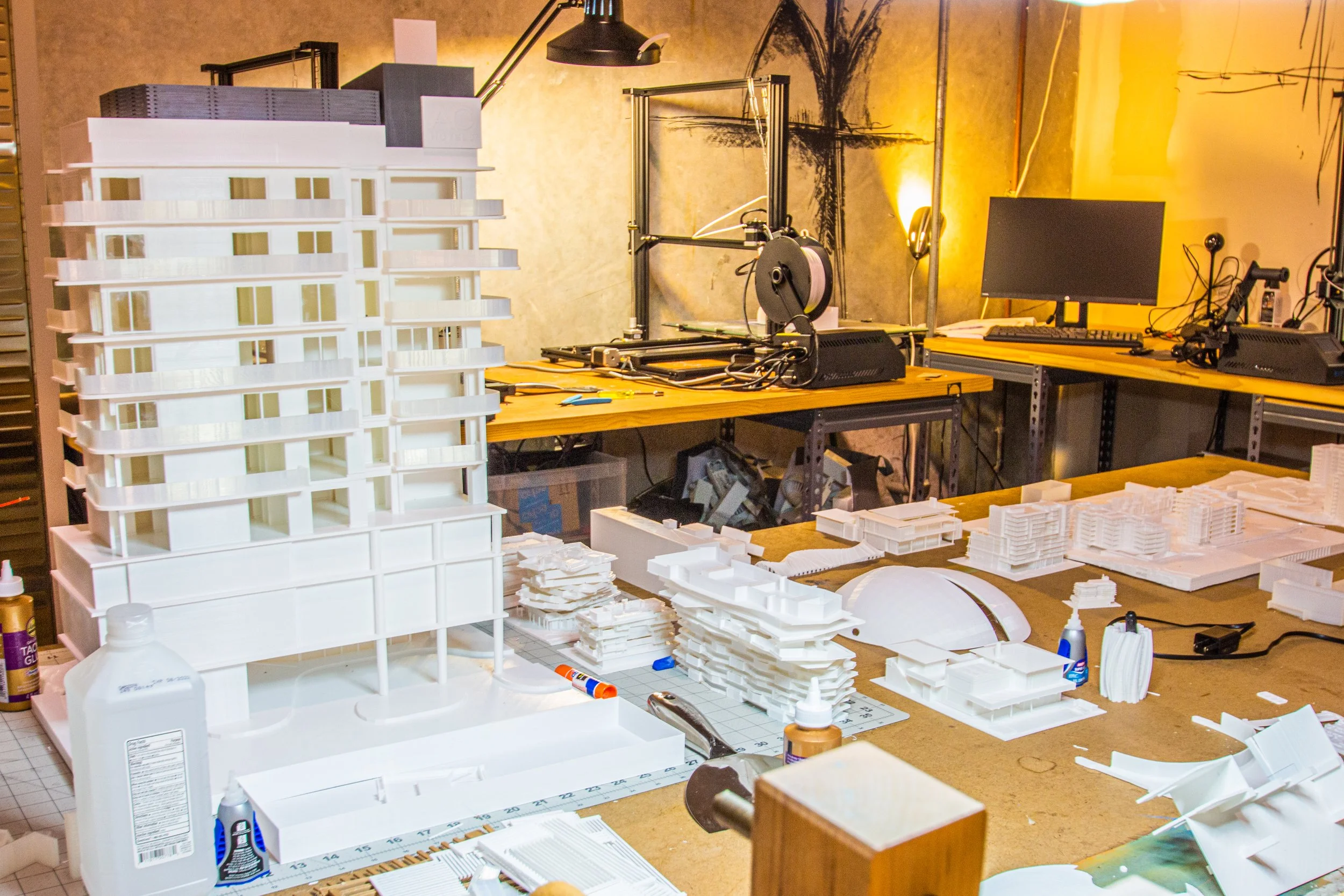 A workspace with various architectural model components, tools, and a 3D printing setup. There is a large white architectural model of a building, workbenches with smaller model pieces, and 3D printers in the background.