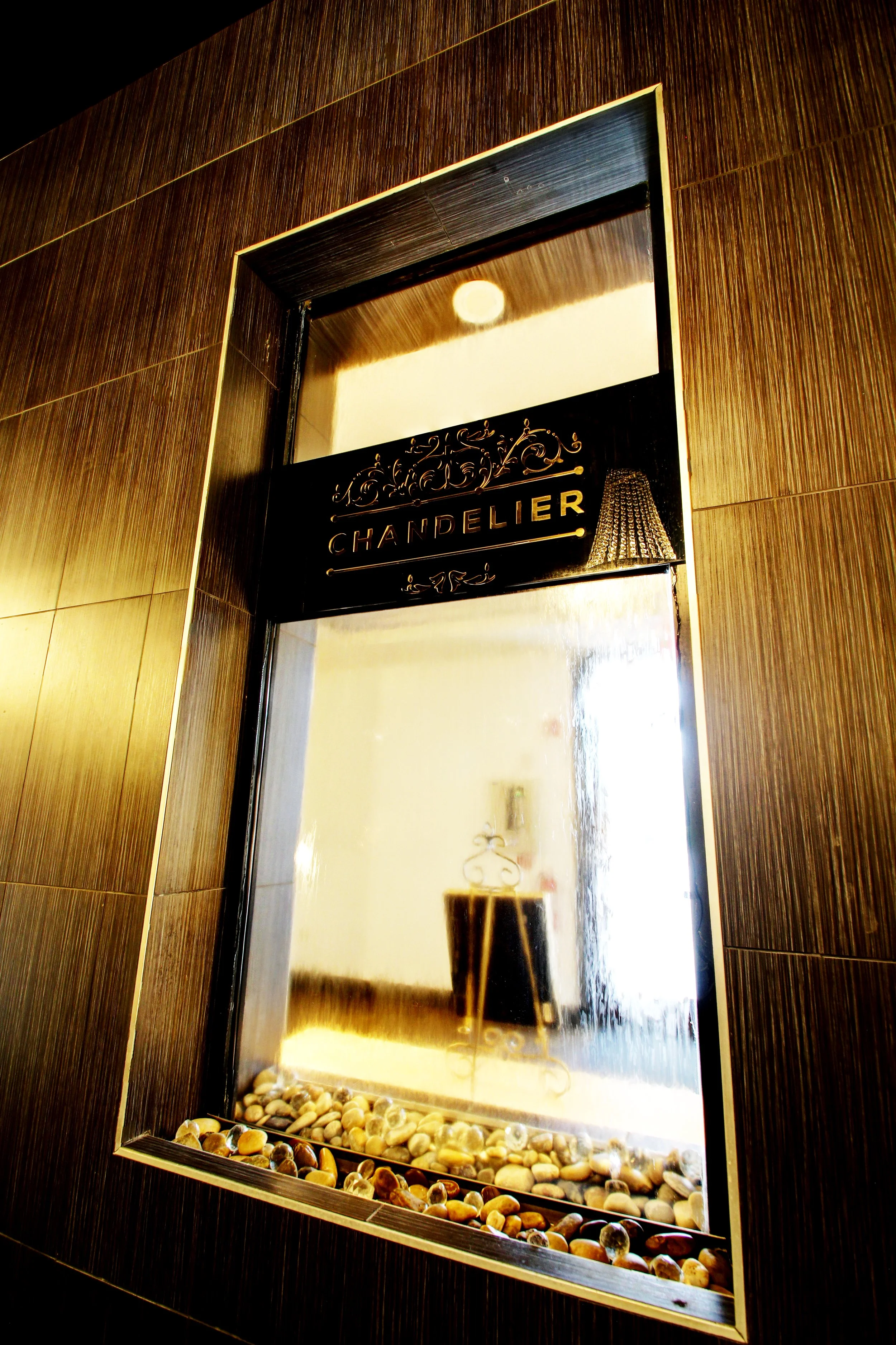 A decorative mirror with a black frame and gold accents, labeled "Chandelier," mounted on a dark wood-paneled wall with pebbles at the bottom, reflecting an interior light fixture and part of a room.