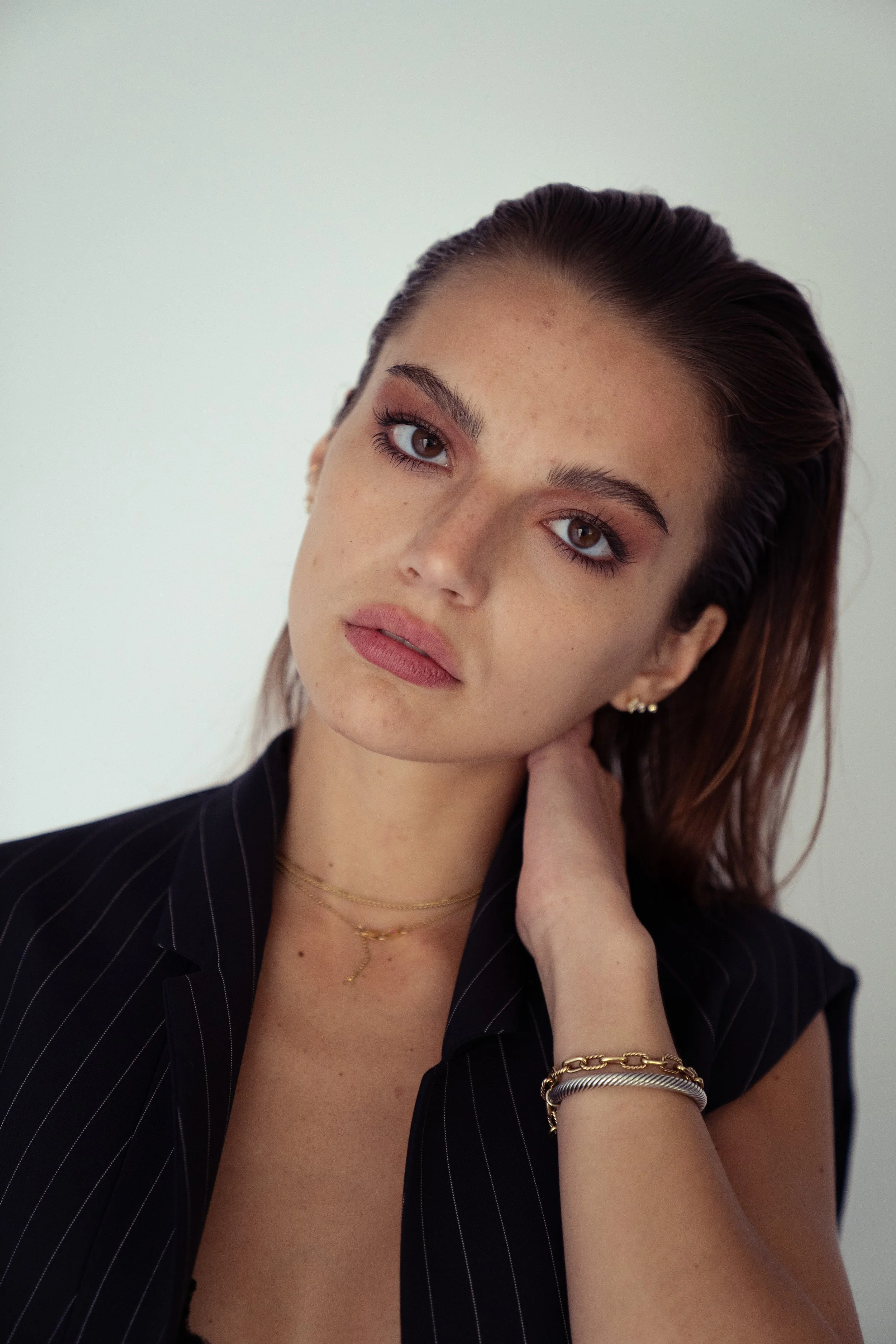 A woman with brown hair, wearing a black pinstripe sleeveless top, gold and silver jewelry, resting her hand on her neck, looking at the camera against a plain background.