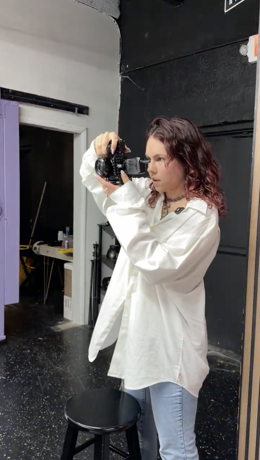 A woman with wavy, reddish-brown hair wearing a white oversized shirt and light blue jeans, holding a camera and taking a photograph in a studio or backstage setting.