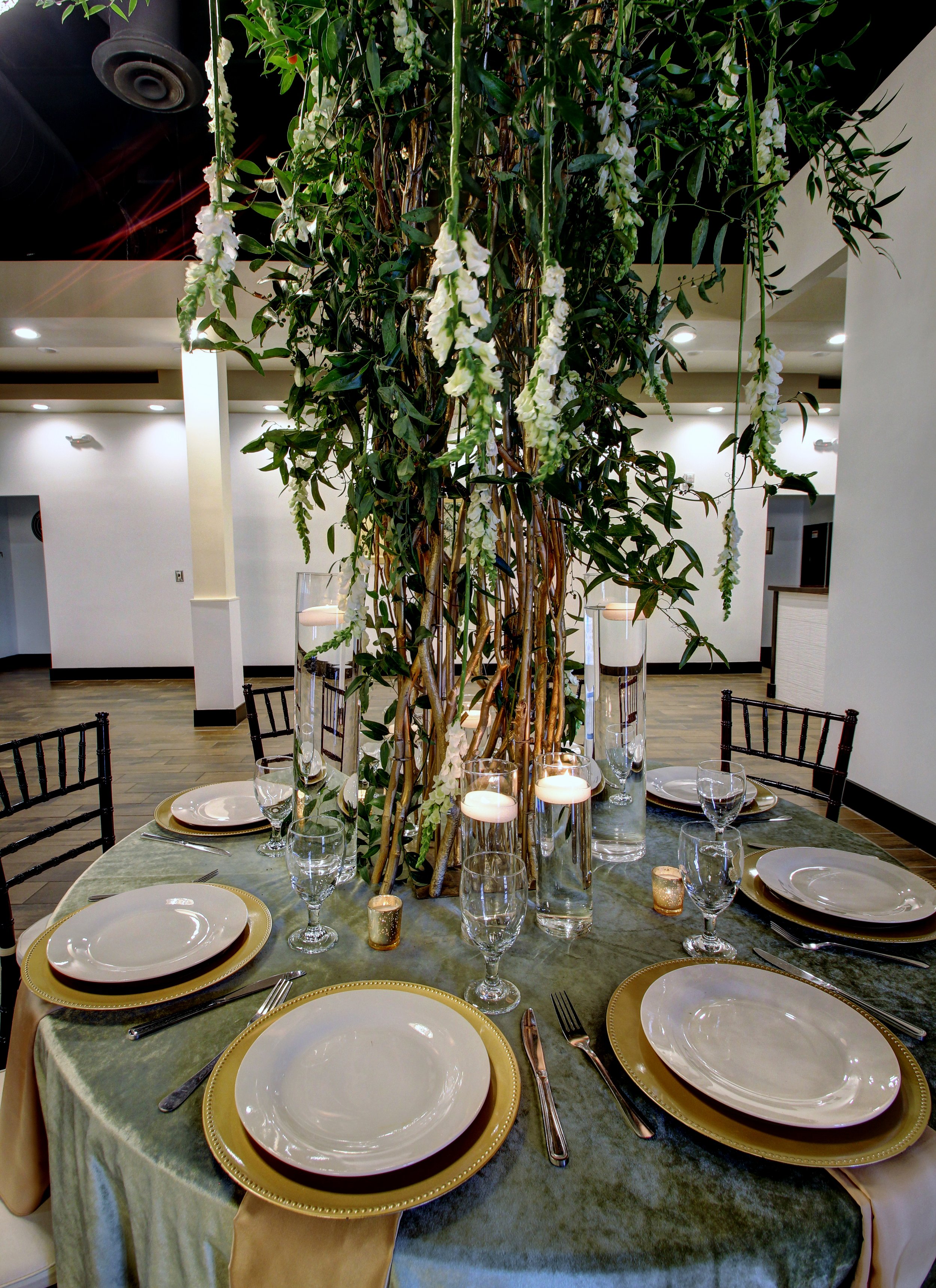 Table setting with white plates on gold-rimmed chargers, silverware, wine glasses, and candles, with a tall floral centerpiece of white flowers and green foliage in a vase, on a round table covered with a green tablecloth in a decorated reception are