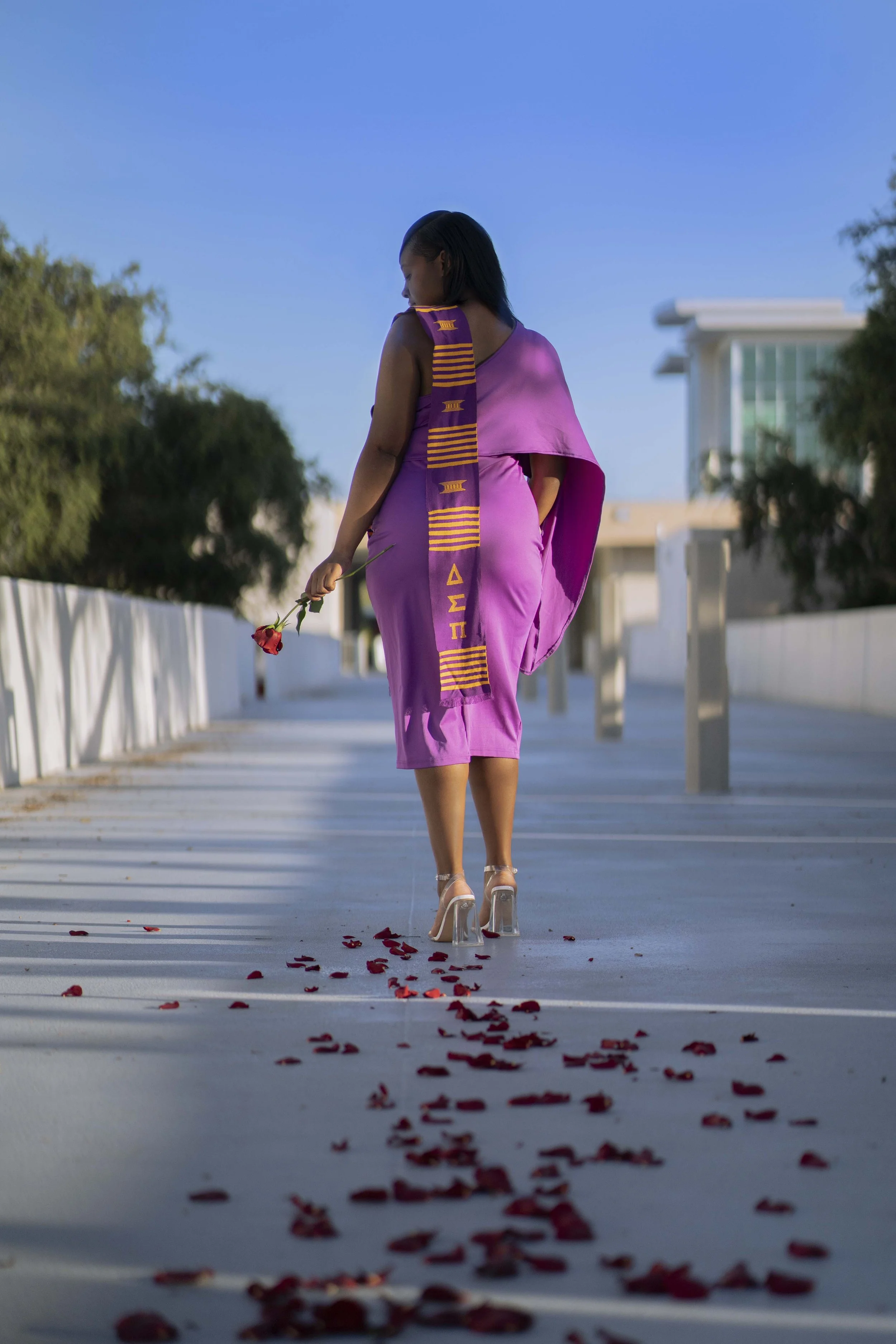 A woman in a purple dress and clear high heels walking on a pathway covered with rose petals, holding a single red rose. She is holding a cloth with HBCU symbols. The pathway is lined with white railings and is in an outdoor setting with trees and mo
