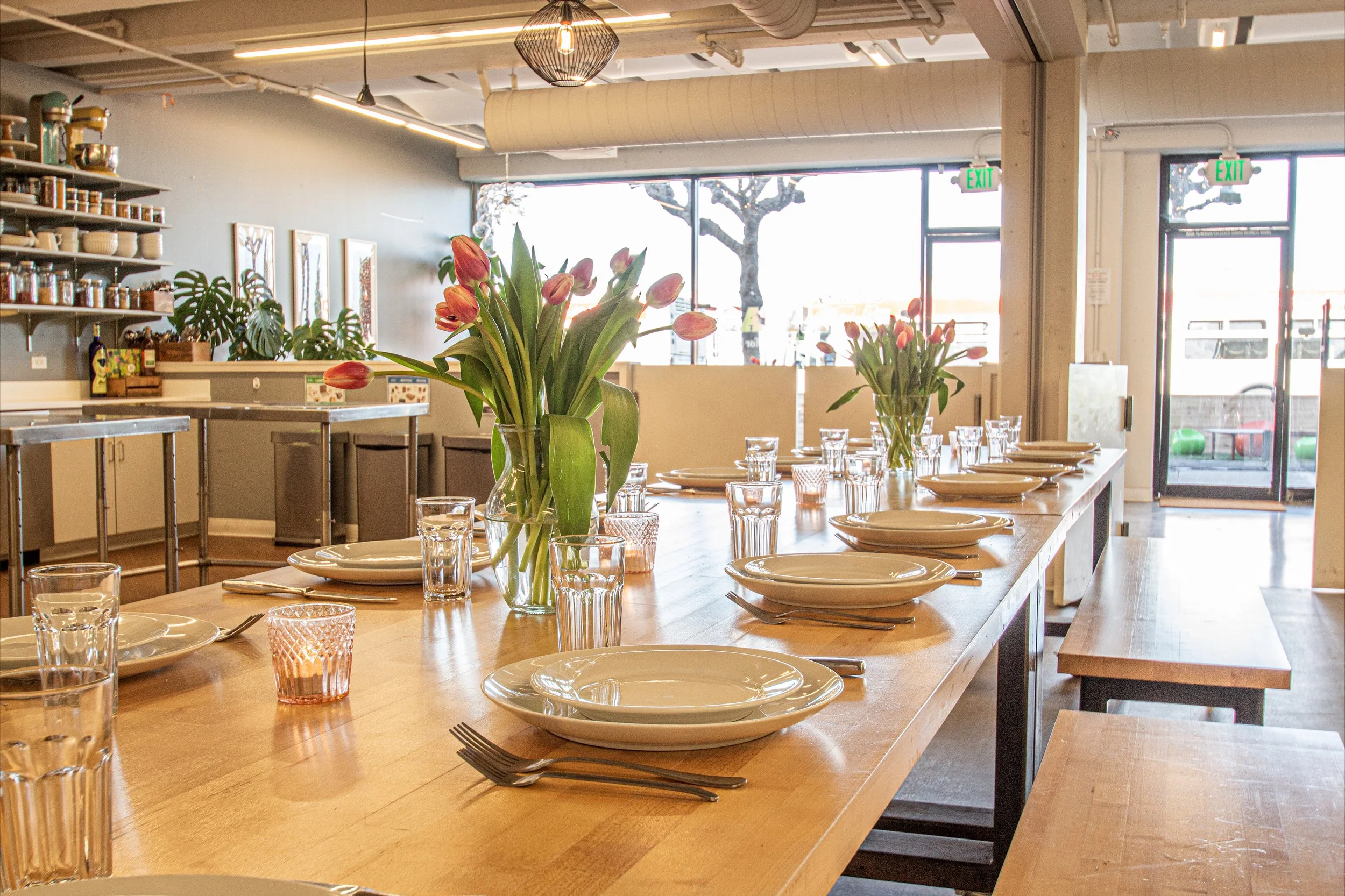 A dining area with a long wooden table set with white plates, forks, glasses, and pink-tinted candle holders. Two vases with pink tulips are in the center of the table. The background features a kitchen area with shelves holding dishes and jars, and 