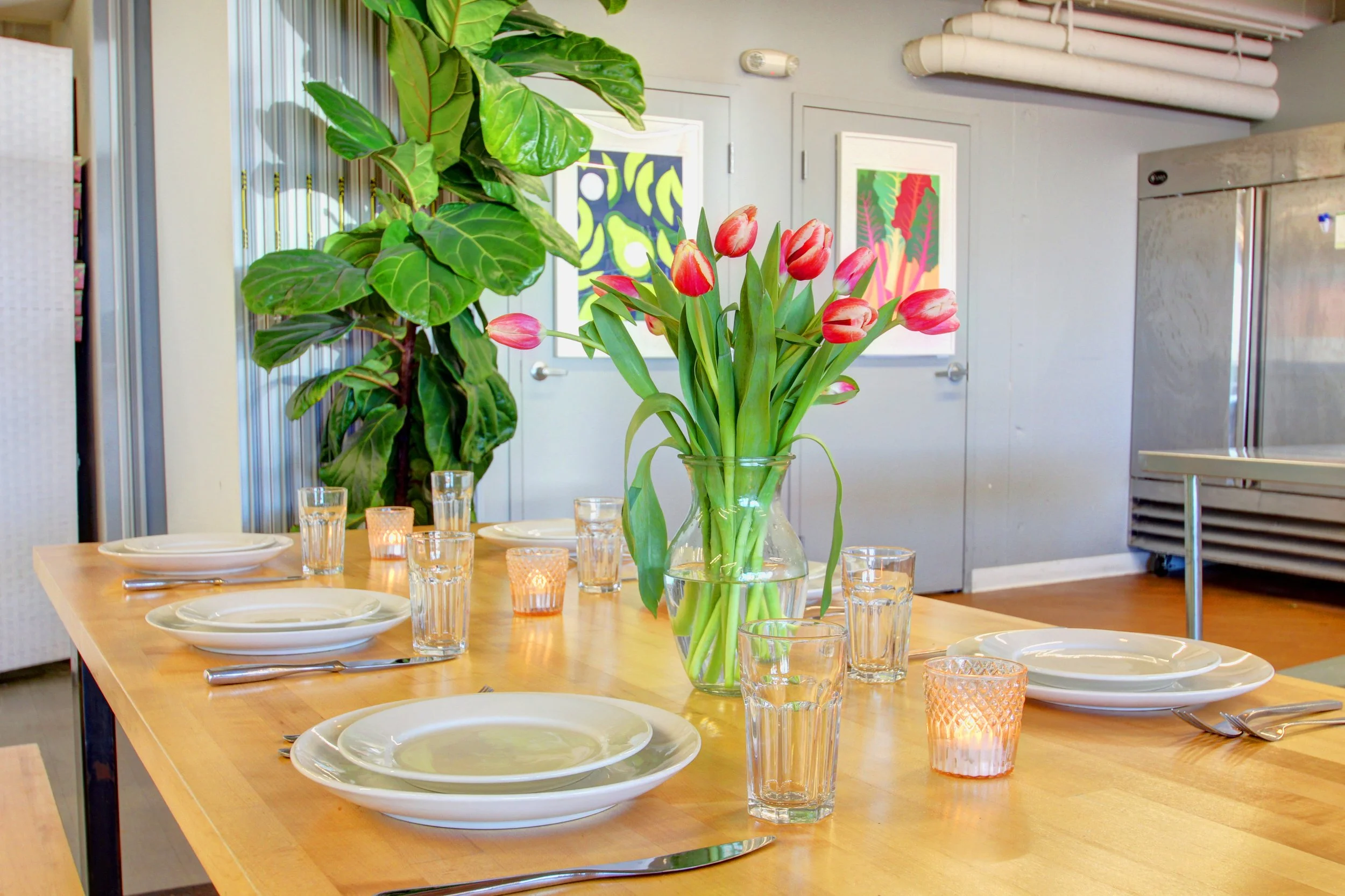 A dining table set with white plates, silverware, clear glasses, small lit candles, and a large glass vase filled with pink tulips. The table is in a room with green plants, colorful wall art, and an industrial-style refrigeration unit.