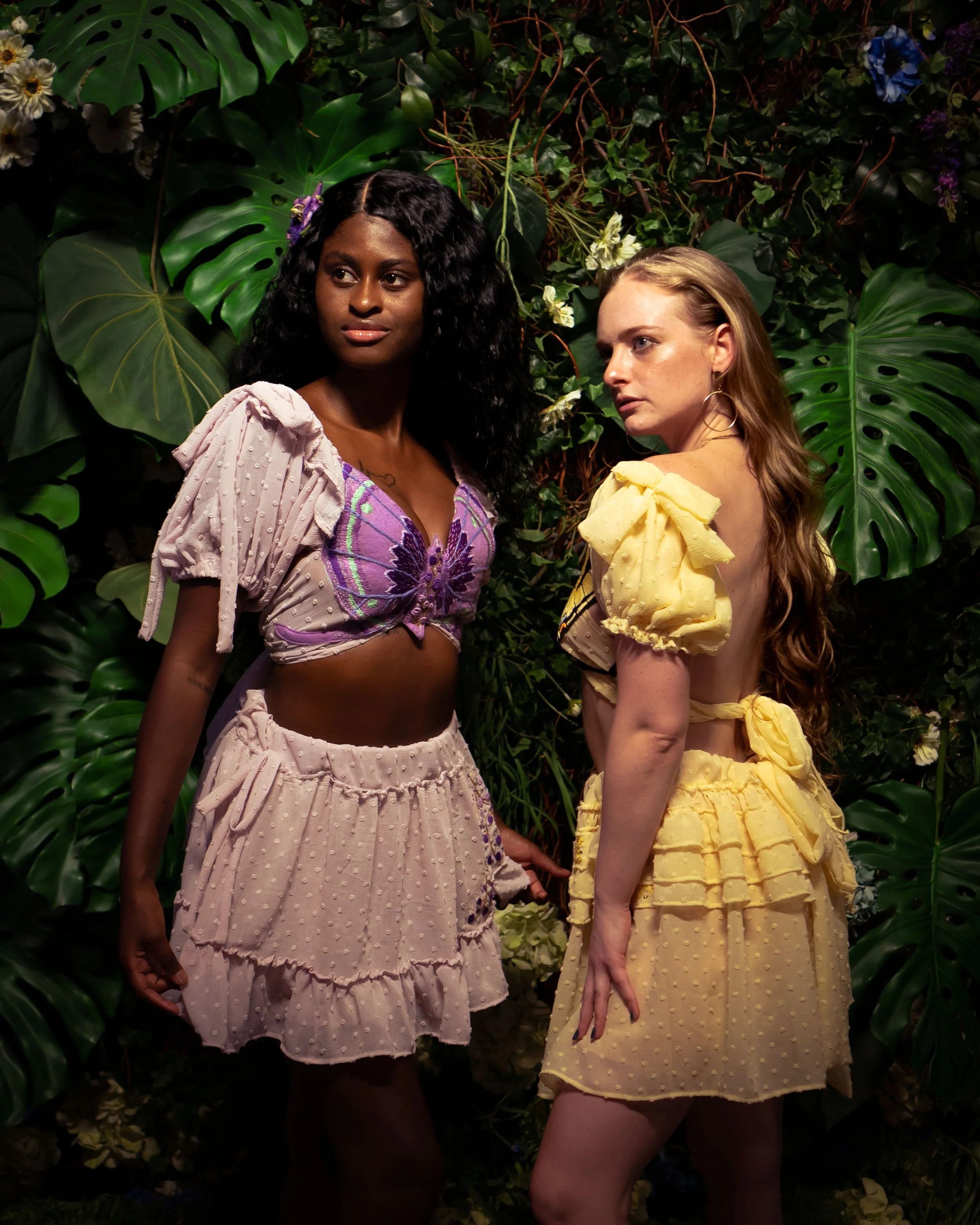 Two women standing in front of a lush green tropical background with large leaves and flowers, wearing colorful, ruffled, puff-sleeve dresses.