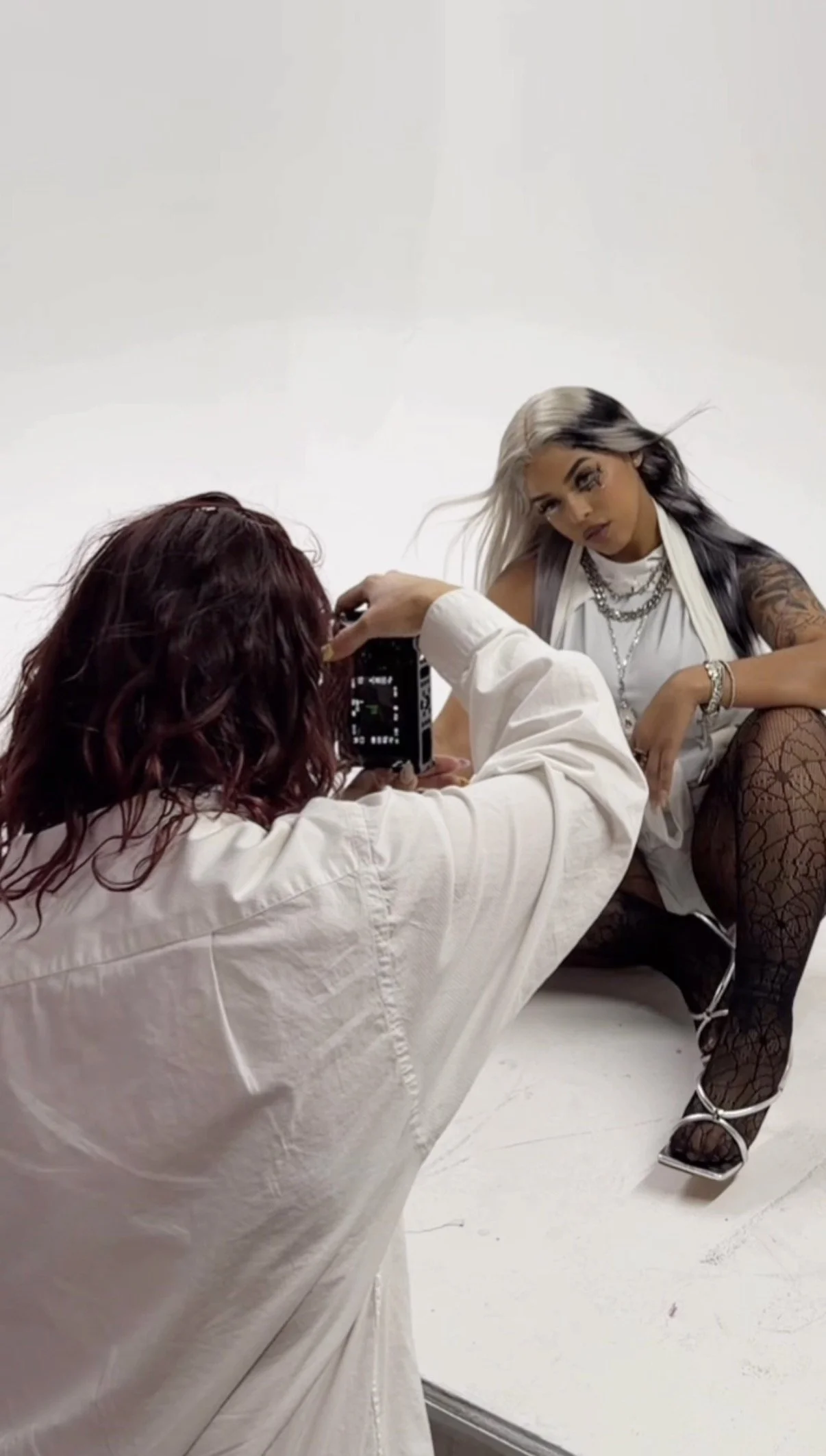 A woman with silver and black hair sitting on a white background, posing for a photo taken by another person with brunette hair and a white shirt, who is holding a camera.