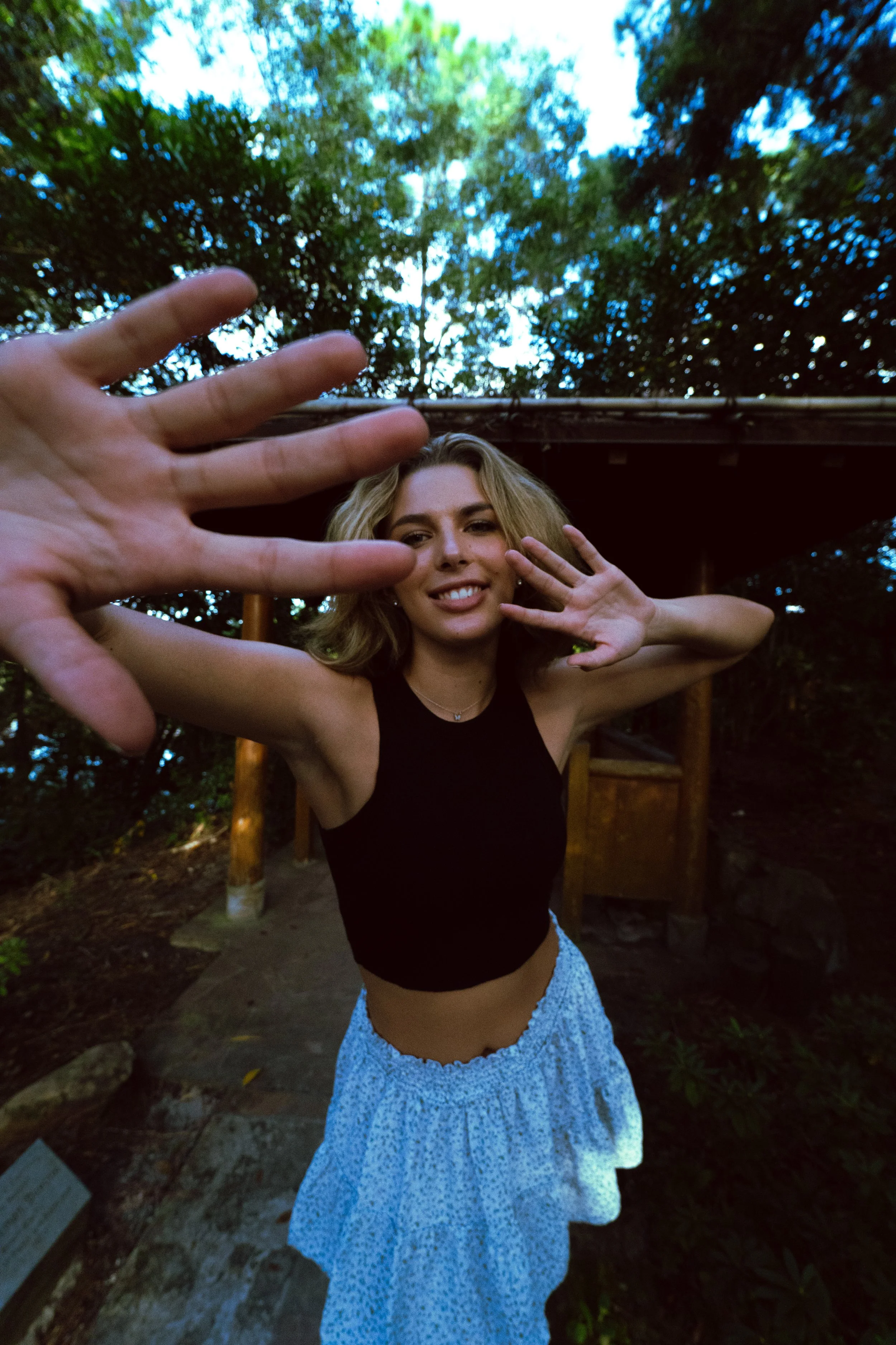 Young woman with blonde hair making a playful gesture towards the camera outdoors, with trees and a wooden structure in the background.