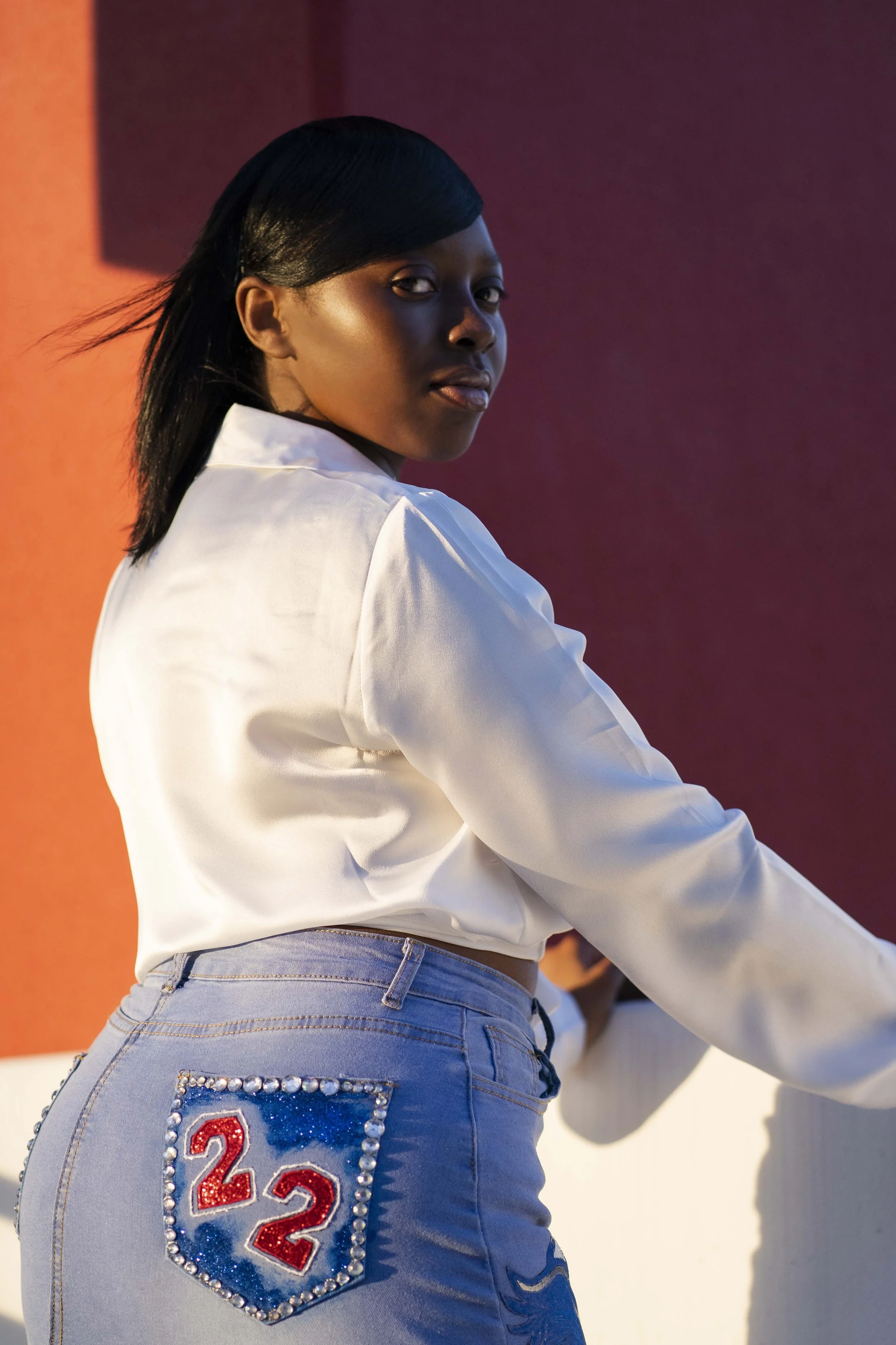 A young woman with black hair streamlined to one side, wearing a white satin blouse and light blue jeans with a rhinestone and glitter patch number 22 on the back pocket, standing outdoors against a colorful background.