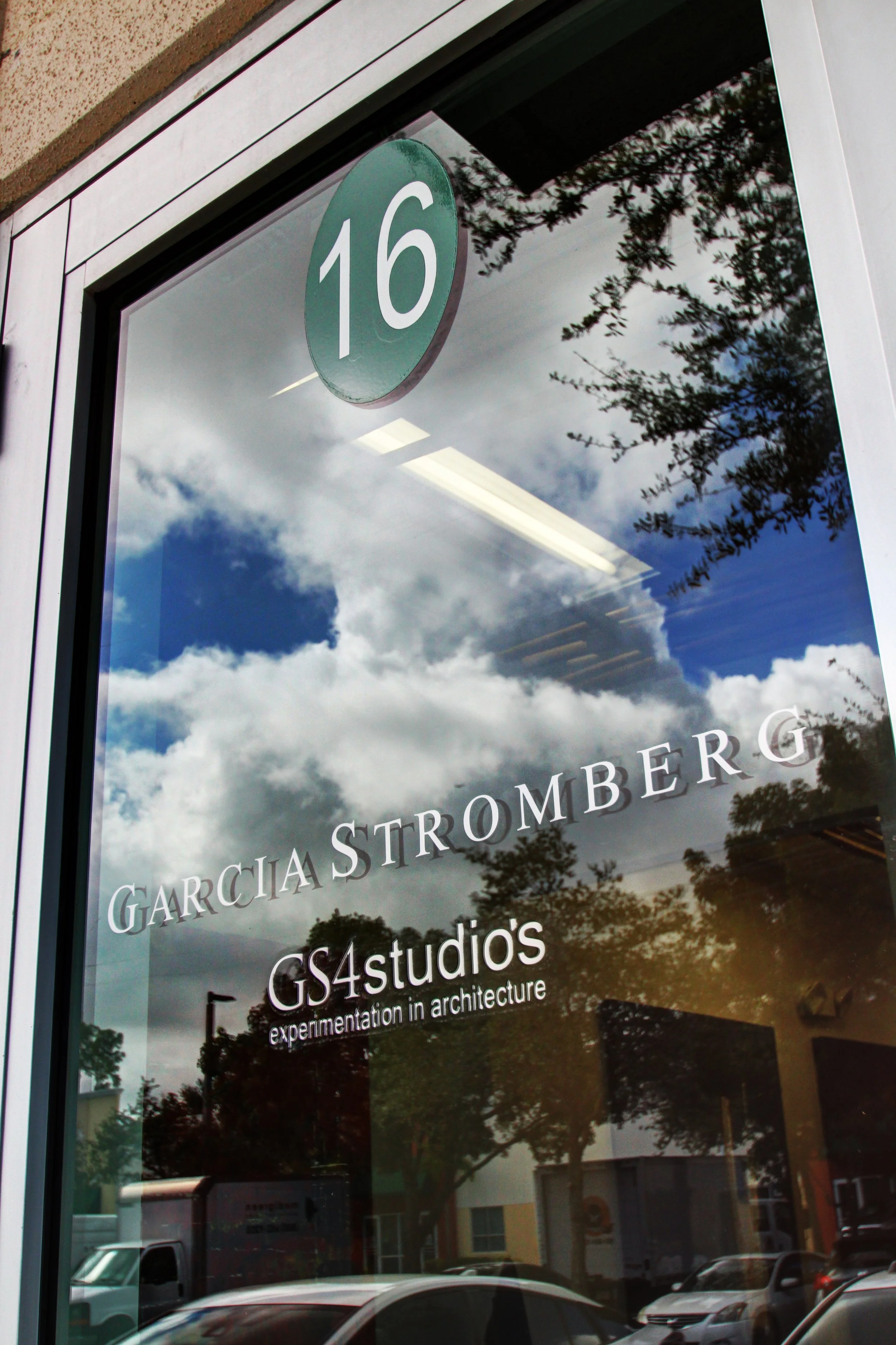 Reflective glass window with the address 16 on a round sign at the top. Text on the window reads Garcia Stromberg GS4studios, experimentation in architecture. The reflection shows a cloudy sky, trees, and parked cars outside.