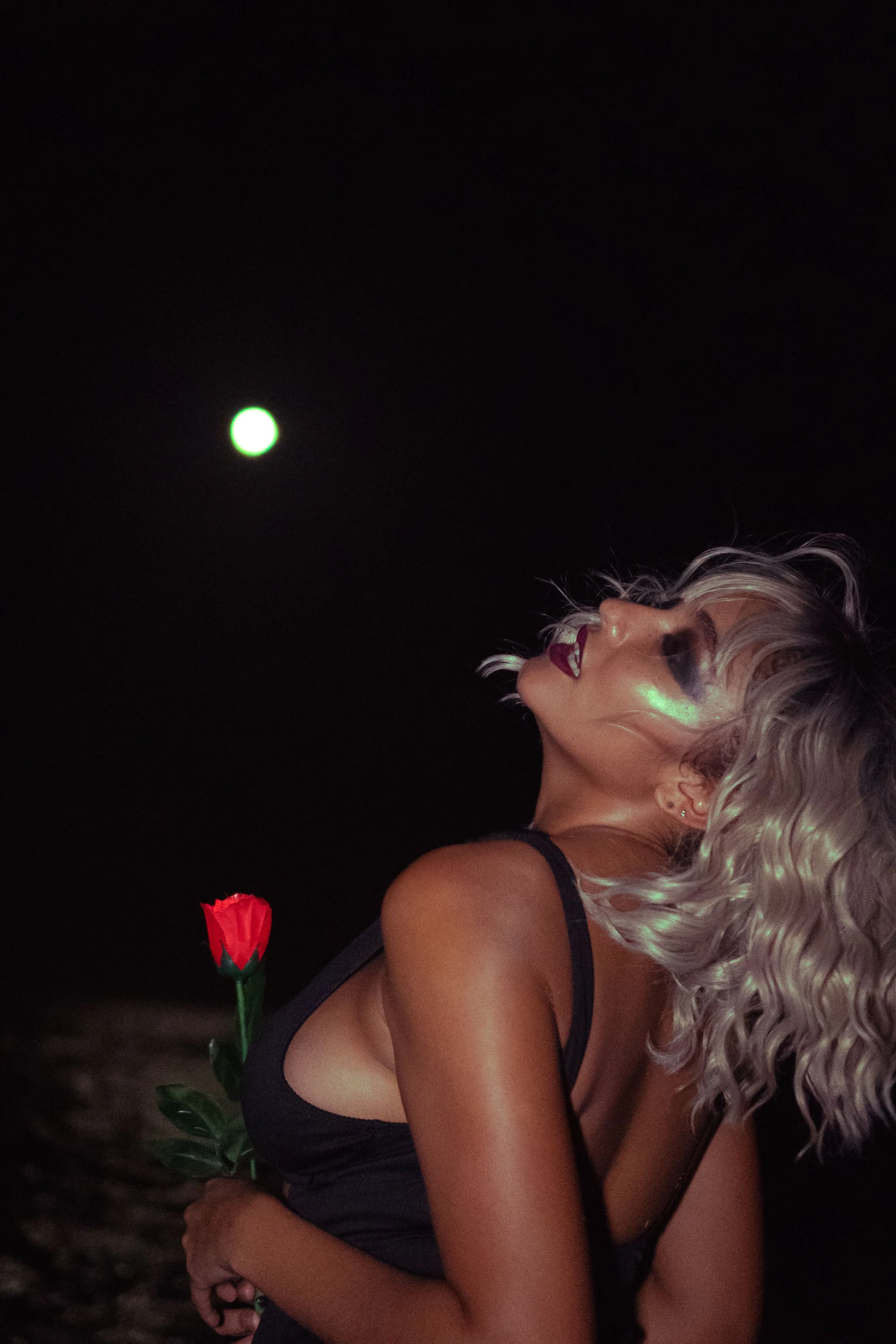A woman with platinum blonde curly hair and dark makeup holding a red rose behind her back at night, with the moon visible in the dark sky.