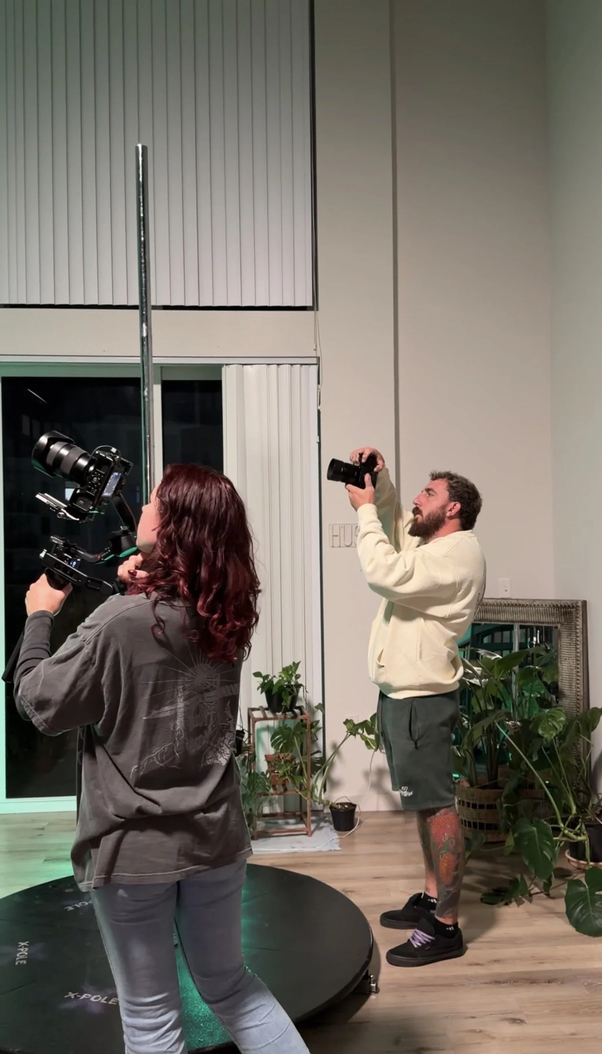 Two people are taking photographs indoors, with various potted plants around them. One person, with long dark hair, is holding a professional camera, and another person, with a beard and tattoos, is holding a camera in the air. They are standing near