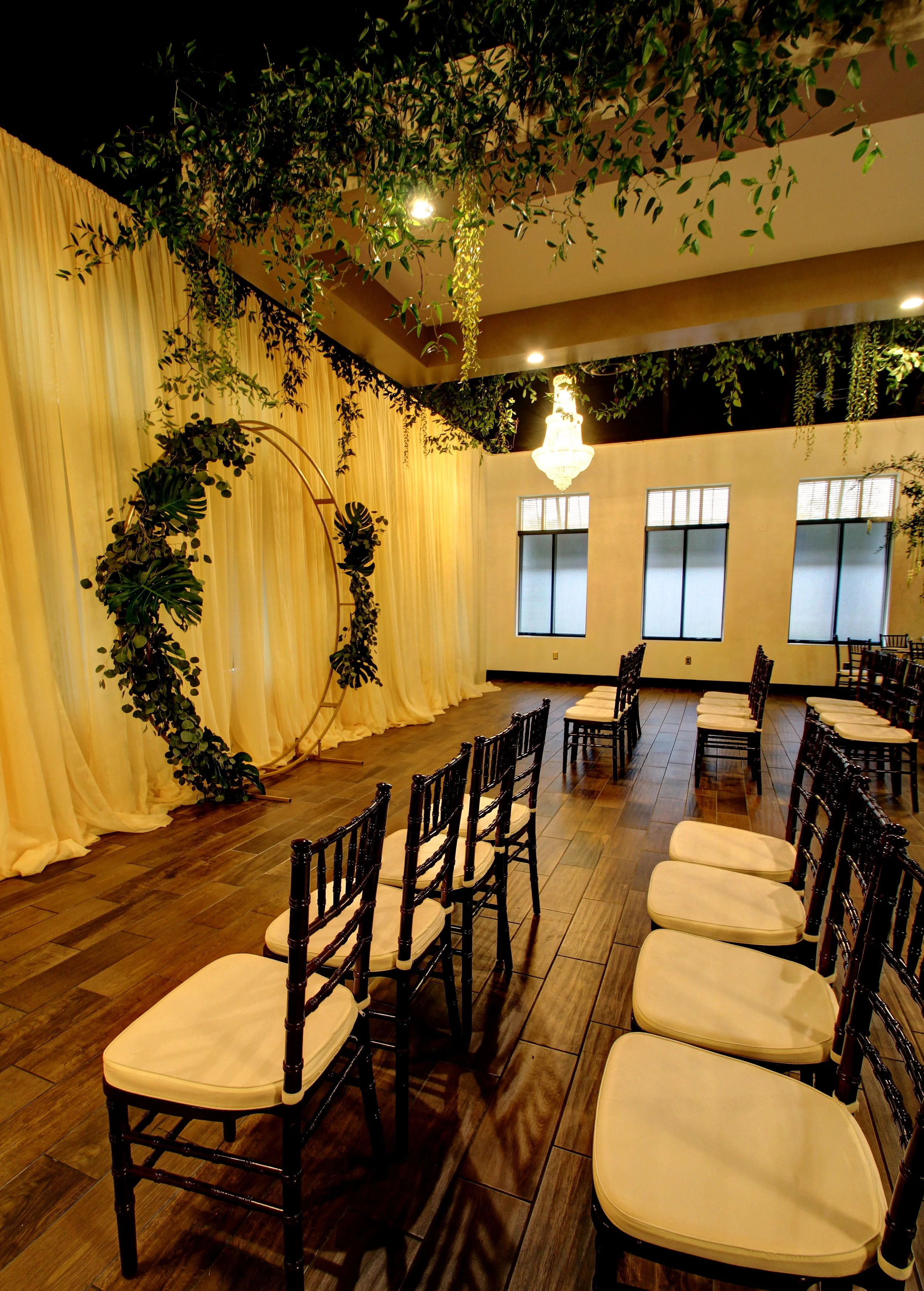 Elegant indoor event space decorated with hanging greenery, a large chandelier, and a circular flower arrangement backdrop on cream-colored curtains, with black chairs with white cushions arranged in rows for seating.