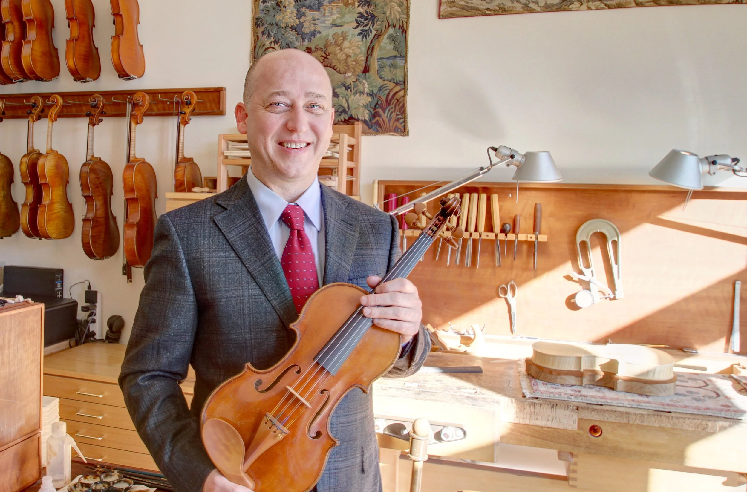 A man in a suit holding a violin in a workshop with violins hanging on the wall and woodworking tools.