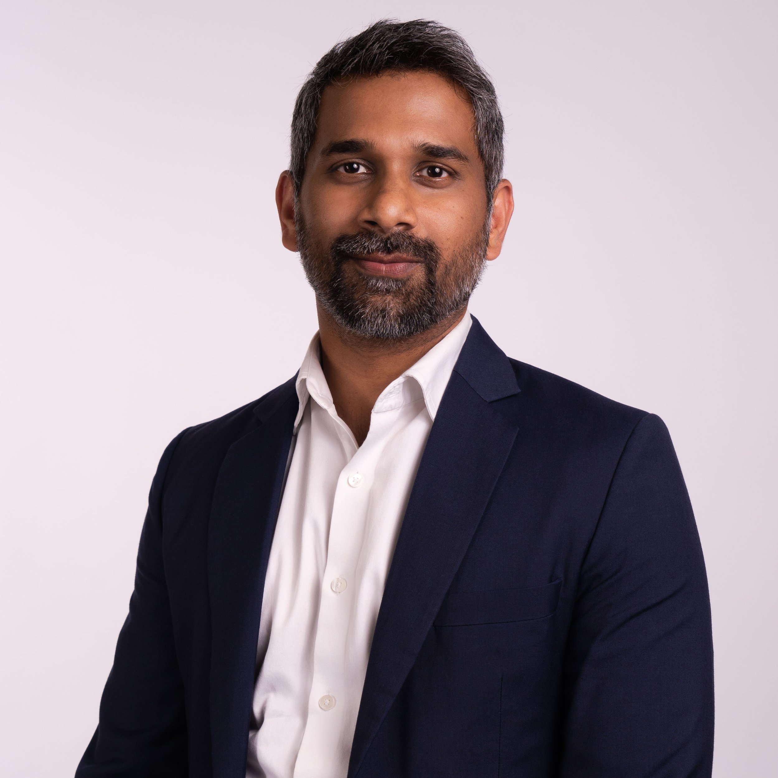 A man with dark hair and a beard, wearing a navy blazer and white shirt, looking at the camera with a neutral expression, against a plain white background.