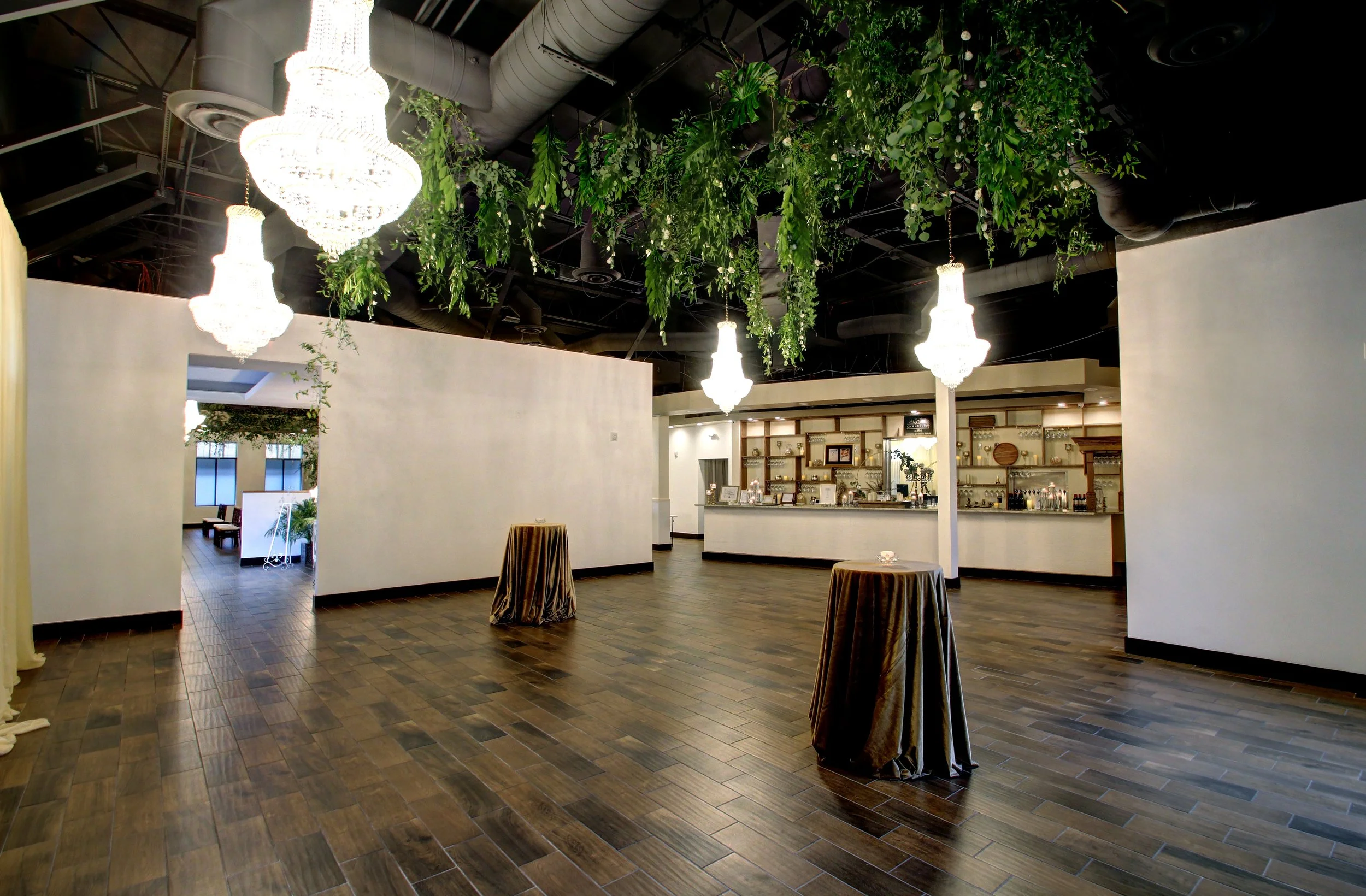 Elegant indoor space with white walls, hanging chandeliers, green hanging plants from the ceiling, and a hardwood floor. There are cocktail tables with draped tablecloths, a bar area, and some furniture visible in the background.