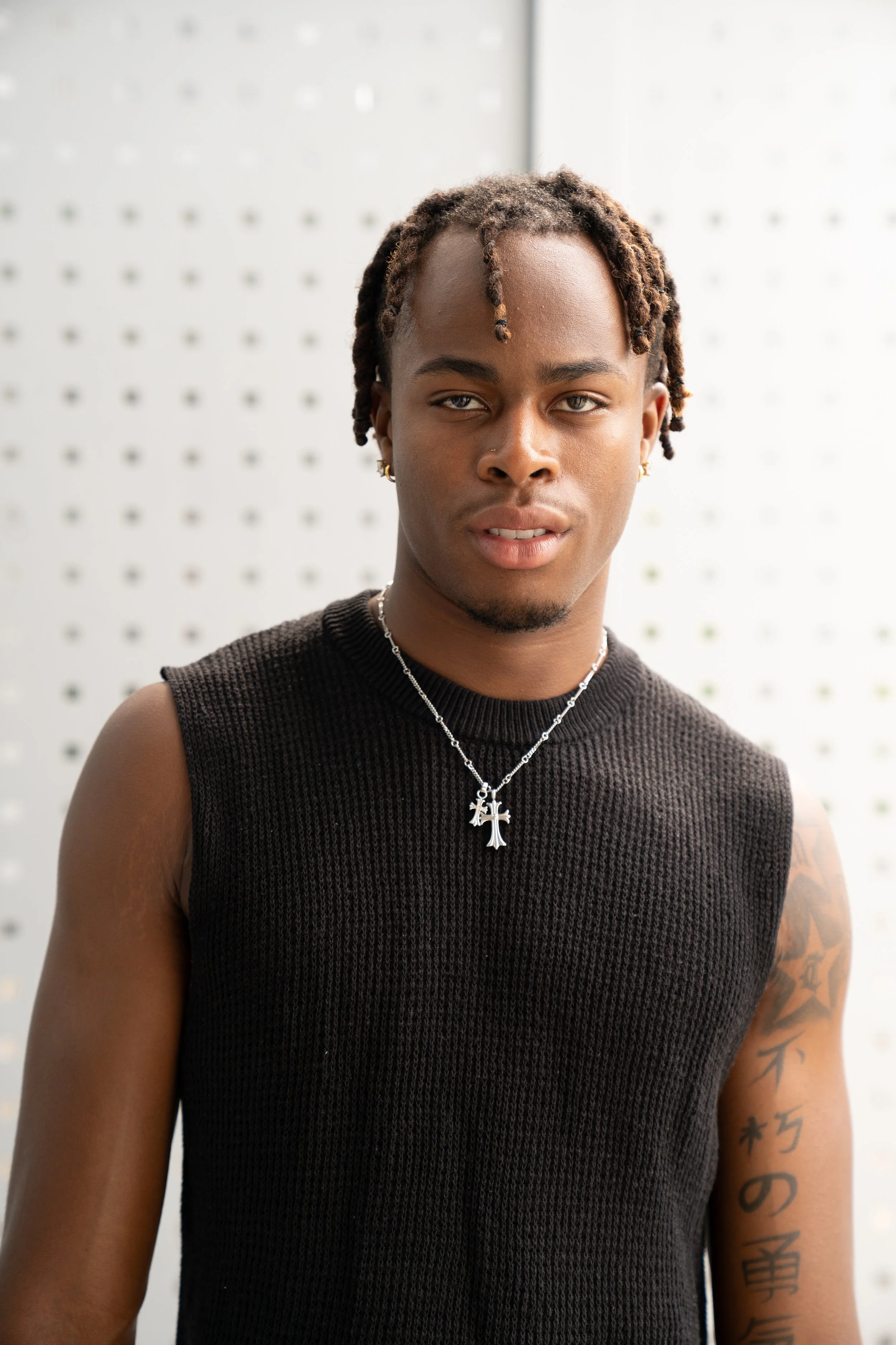 A young man with dreadlocks wearing a sleeveless black knit shirt, silver cross necklace, and earrings, standing in front of a white pegboard wall.
