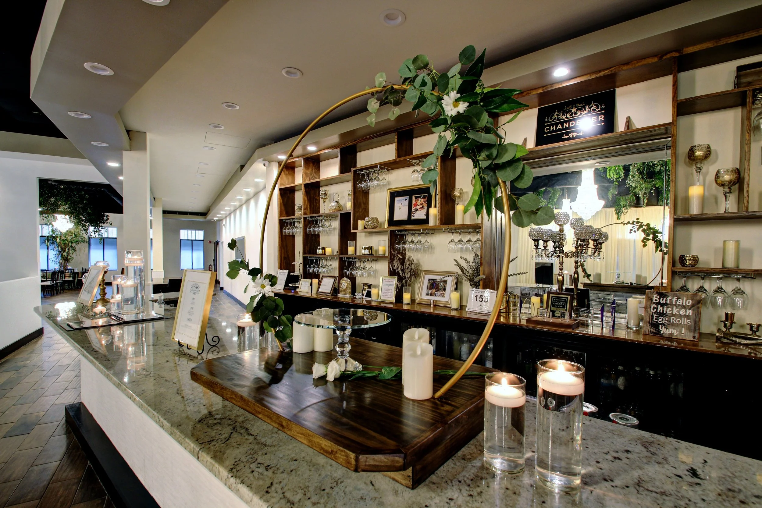 Interior of a restaurant bar area decorated with a floral frame centerpiece, candles, and framed pictures on the counter, with shelves holding glassware and decor, and large windows in the background.