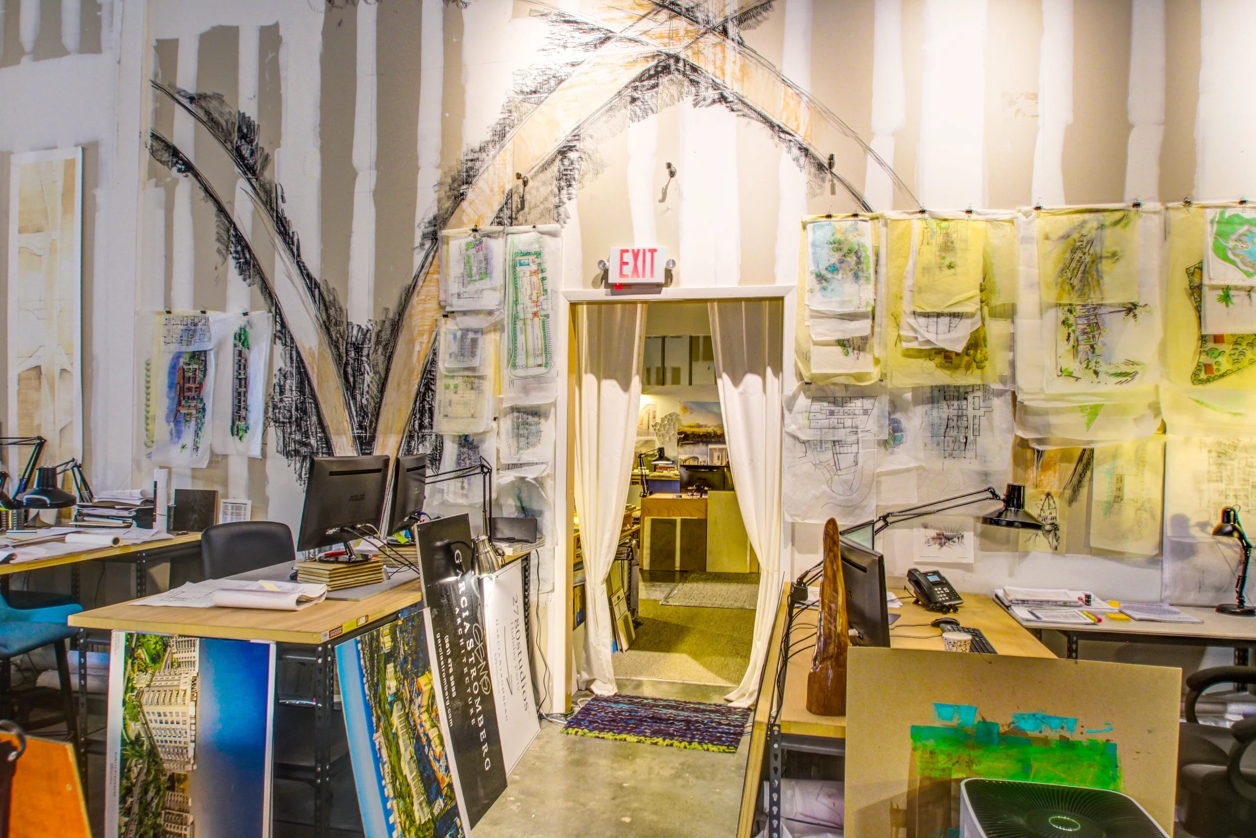 An office or studio space with multiple desks, computers, and art supplies. The wall features artistic sketches and blueprints, with a doorway curtain leading to another room.