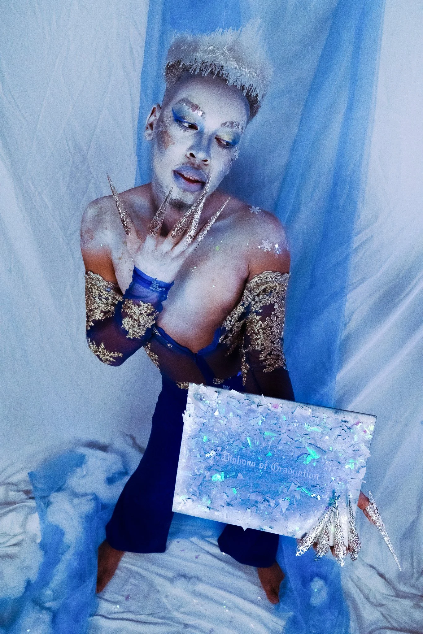 Person with elaborate makeup and costume holding a gift box, surrounded by a blue and white setting with fantasy elements.