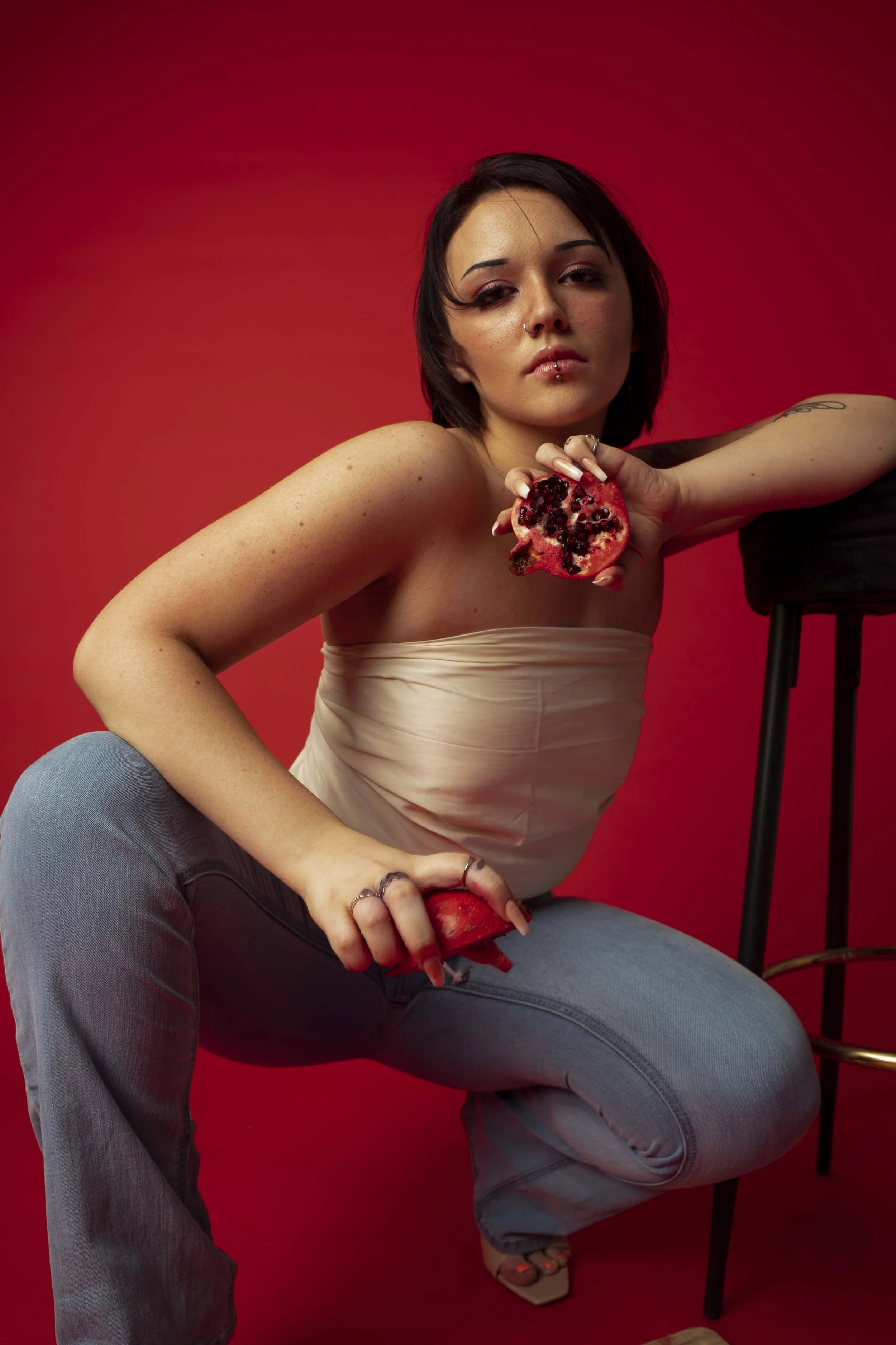 A woman with dark hair, wearing a strapless cream top and jeans, poses against a red background, holding a partially eaten pomegranate in one hand and a section of the fruit in her lap.