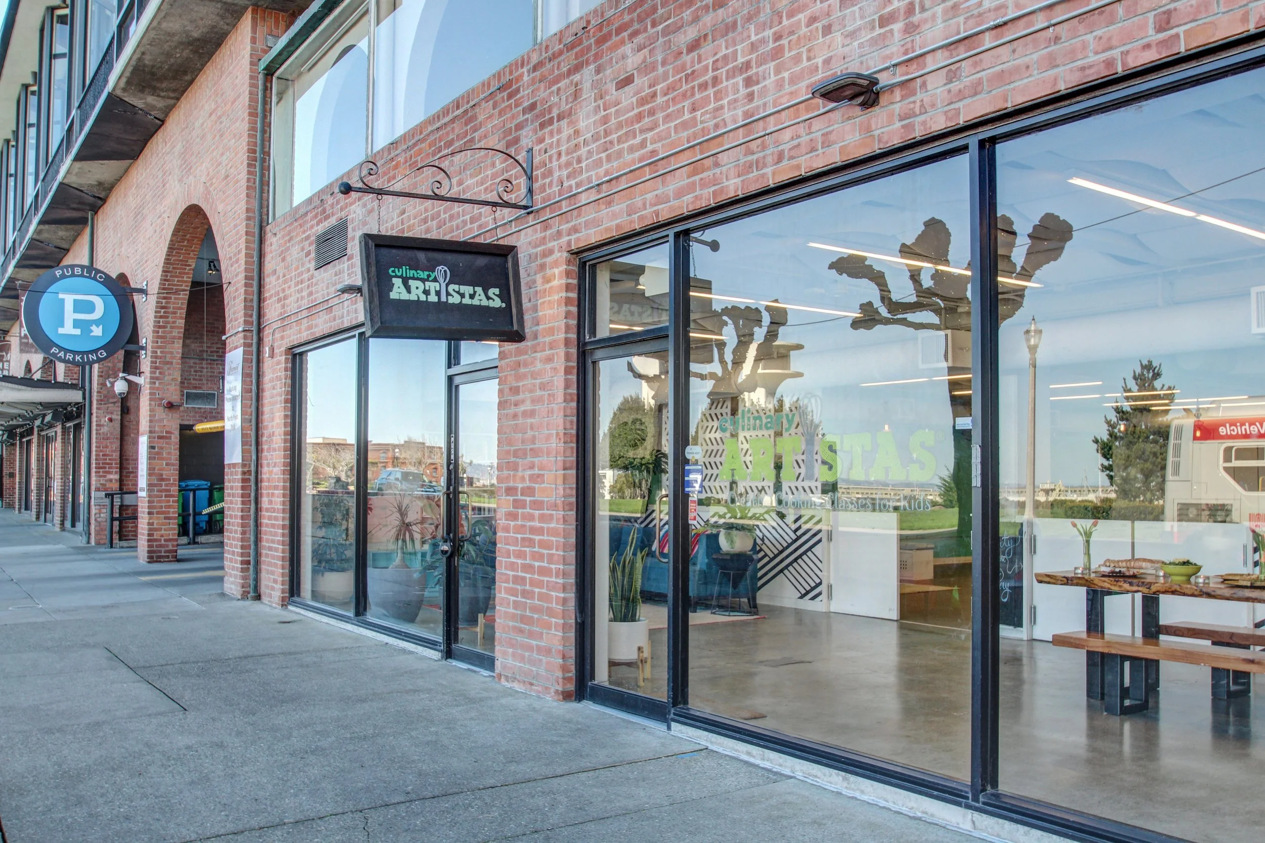 Storefront with large glass windows and a sign that reads "culinary ARTISTAS" on the brick building. Reflects a tree and sky in the glass.