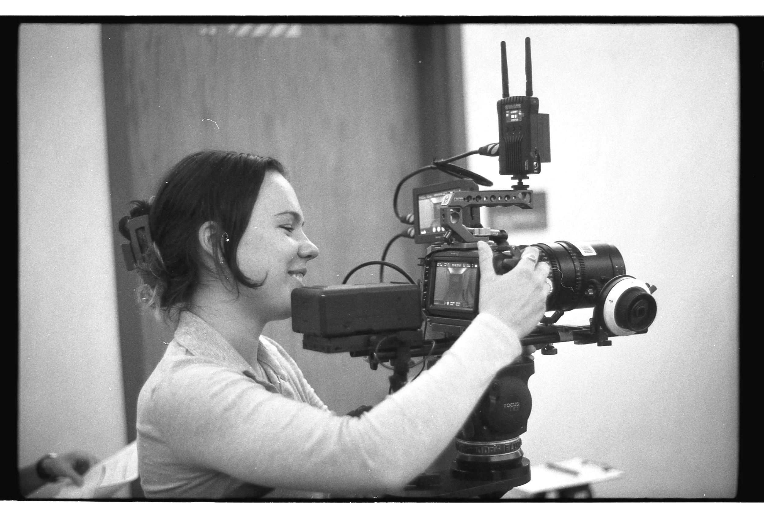 A woman operating a professional video camera with a large lens, on a tripod, smiling as she looks through the camera viewfinder in a studio setting.