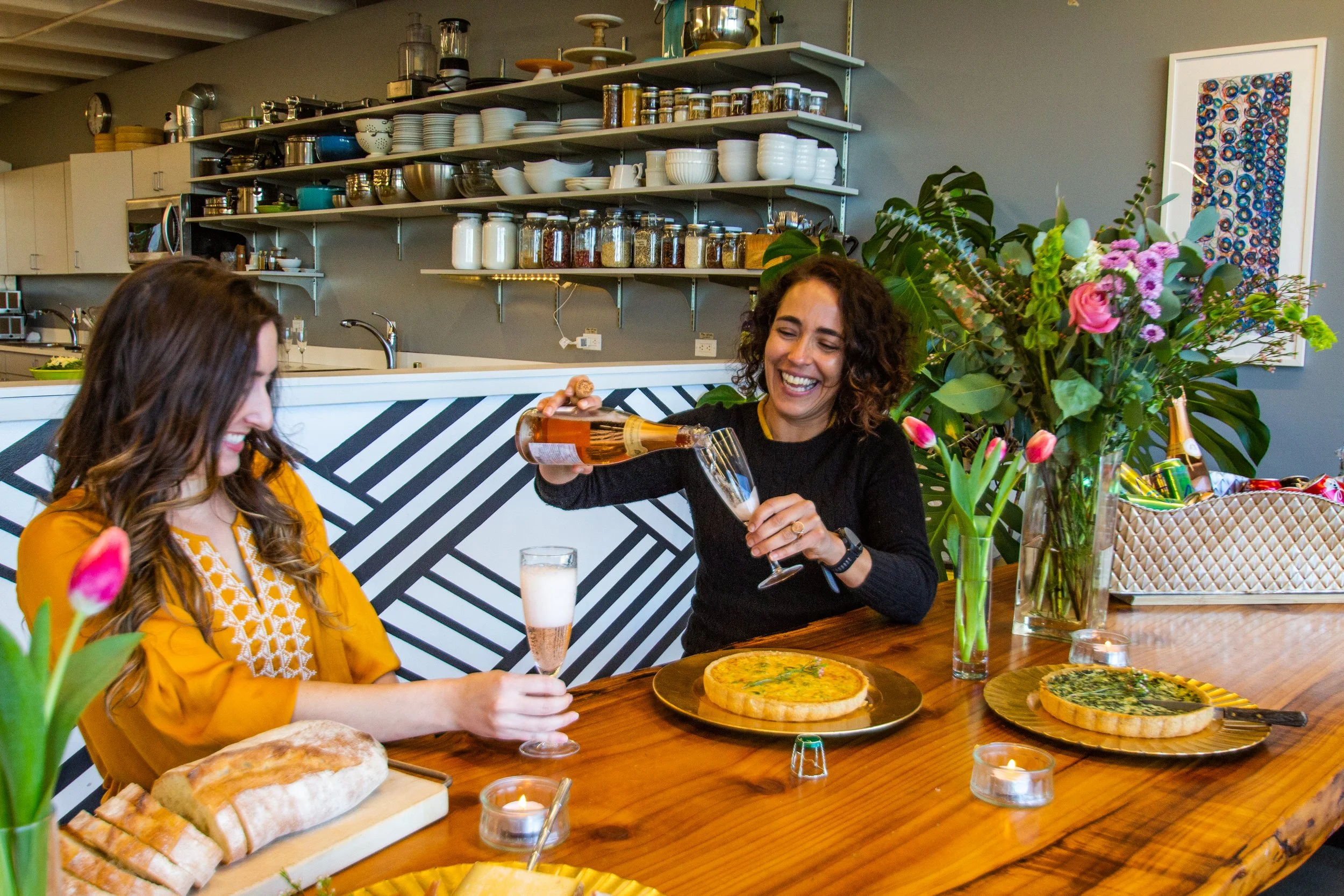 Two women enjoying a meal and drinks at a wooden table in a cozy, well-decorated kitchen or café setting. One woman is pouring rosé wine into a glass while the other woman is holding a filled glass, with a pizza, bread, and flowers on the table.