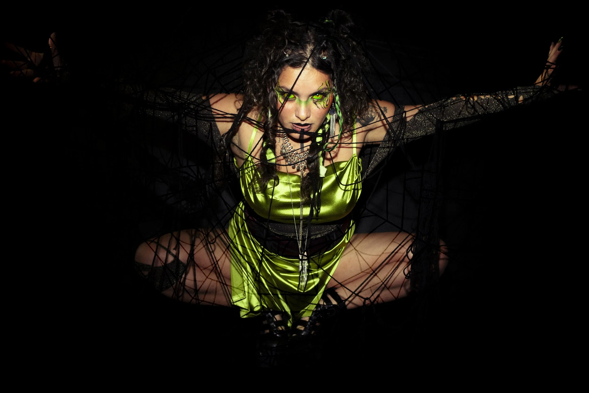 Woman with striking green makeup and hair accessories, dressed in lime green satin outfit, kneeling with arms outstretched behind a web-like structure against a black background.
